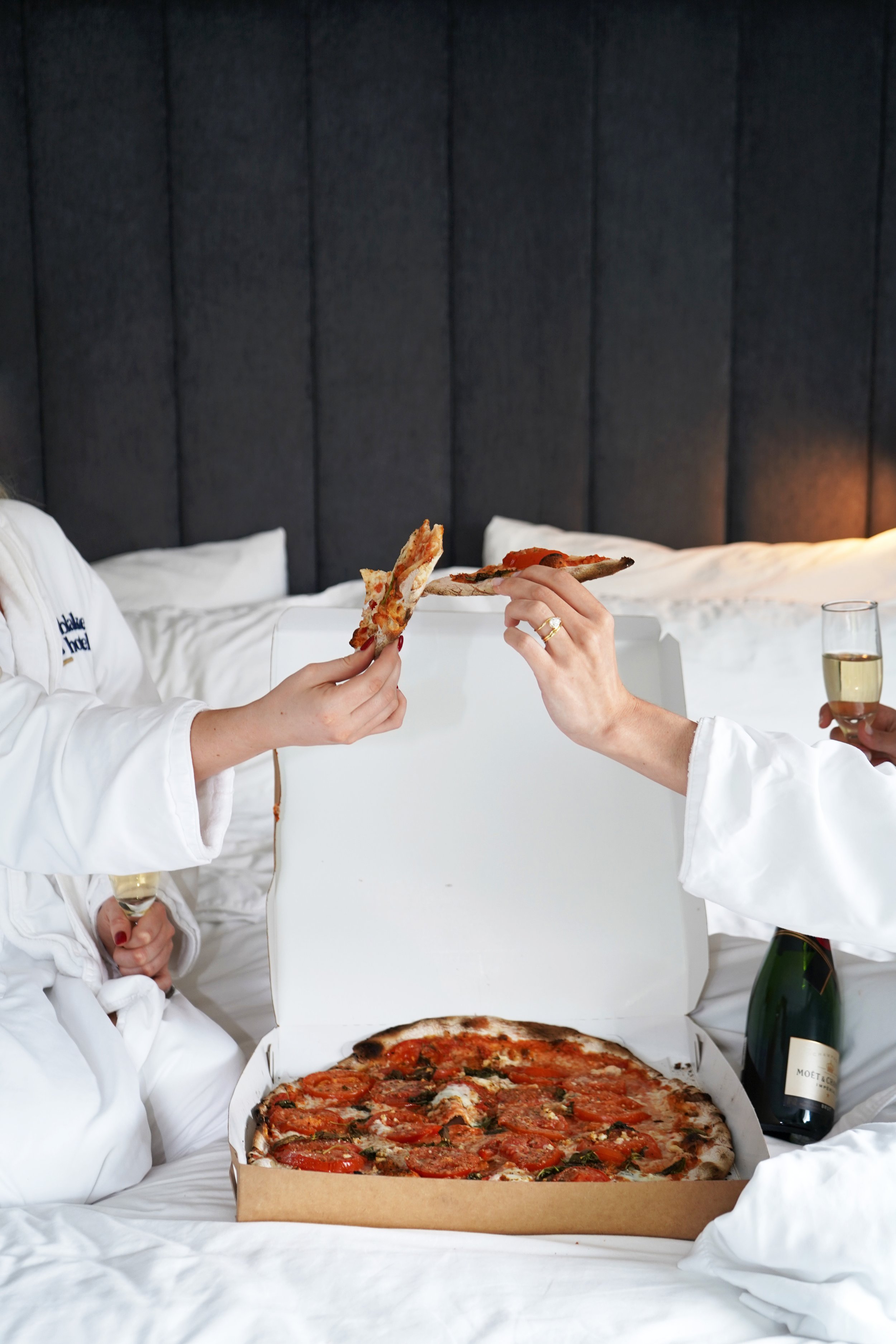 People in white robes sharing pizza and champagne in bed.