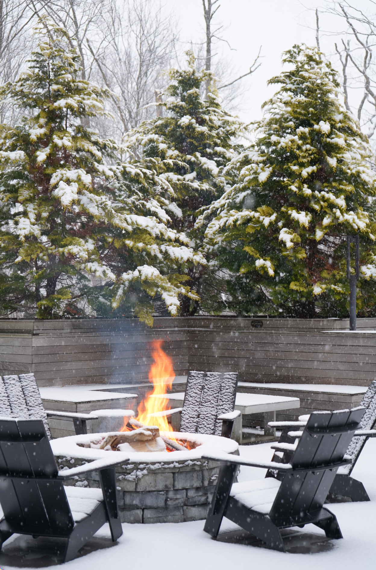 Snow-covered outdoor patio with a burning fire pit surrounded by black chairs, with snow-covered trees in the background.