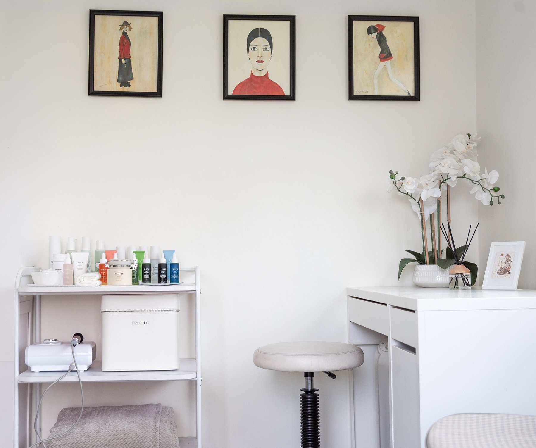 A minimalist interior with three framed art prints on white wall, white furniture, laundry shelf with bottles, a small white cabinet, a stool, a potted orchid, and a framed photo.