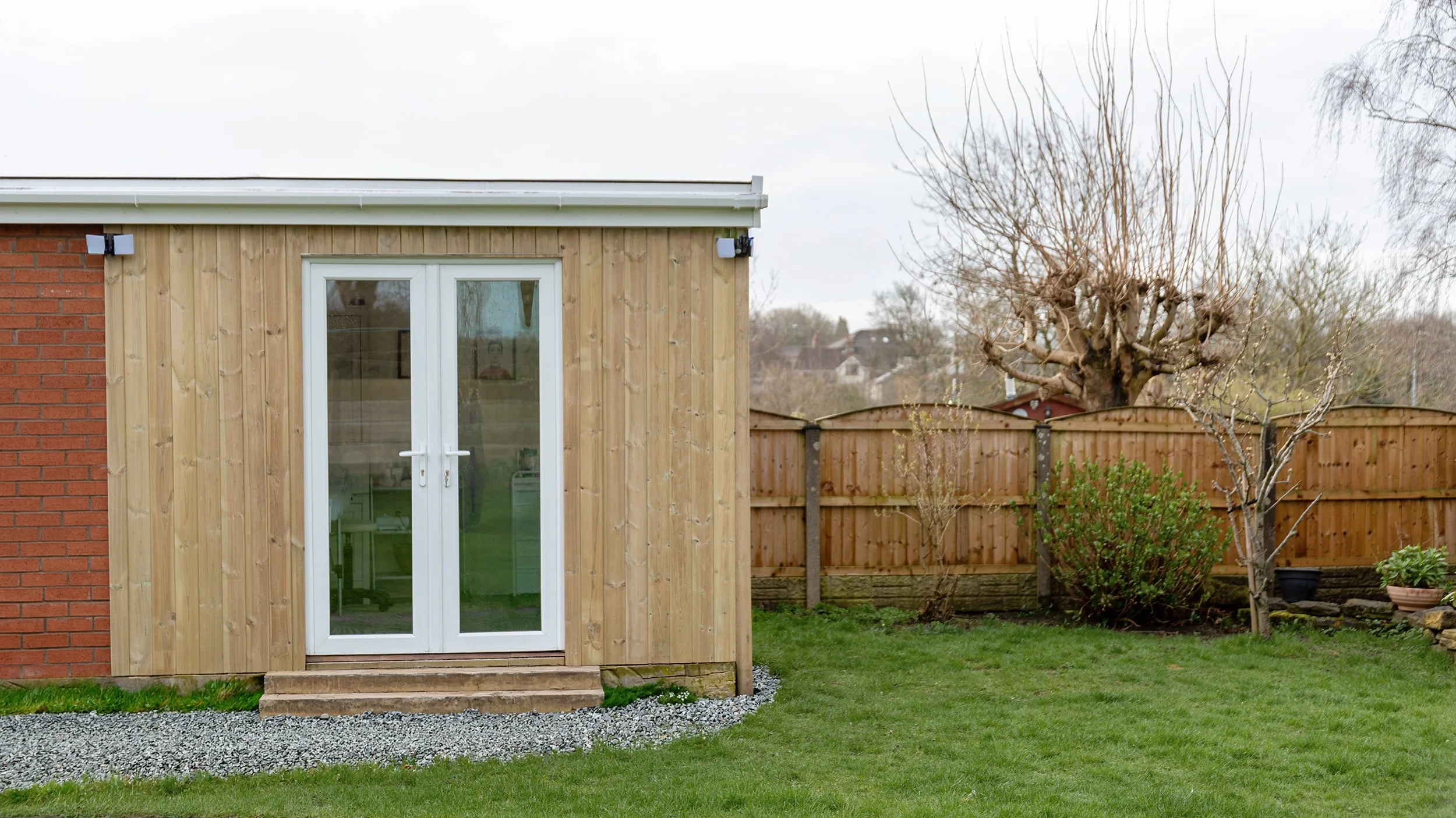 A backyard with a wooden shed that has a glass door, a lawn, and a wooden fence. There are trees and potted plants in the background.