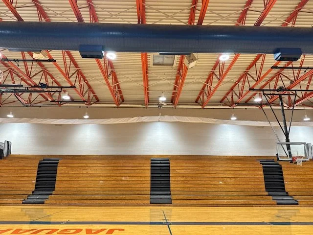 Gymnasium with wooden bleachers and a basketball hoop on the right side with archery curtain above.
