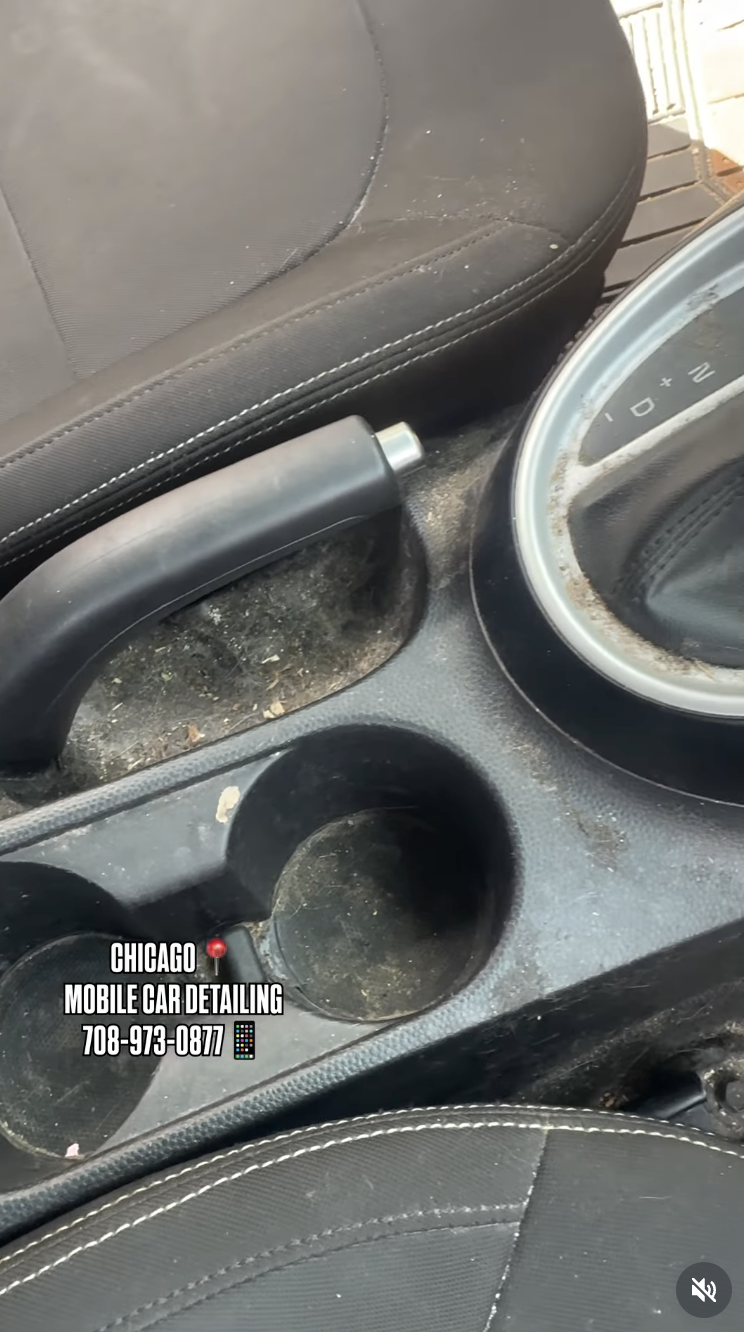 A car's interior with a black center console that has dirt and debris, a speedometer showing 0, a parking brake handle, and contact information for Chicago mobile car detailing.