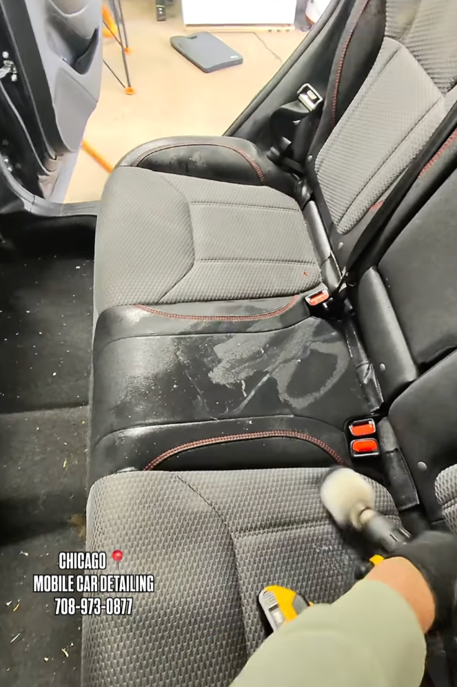 Interior of a vehicle showing the back seats being cleaned with a handheld cleaning tool, with dirt and stains visible on the black surface, and a cleaning mat or tool on the floor near the front of the vehicle.