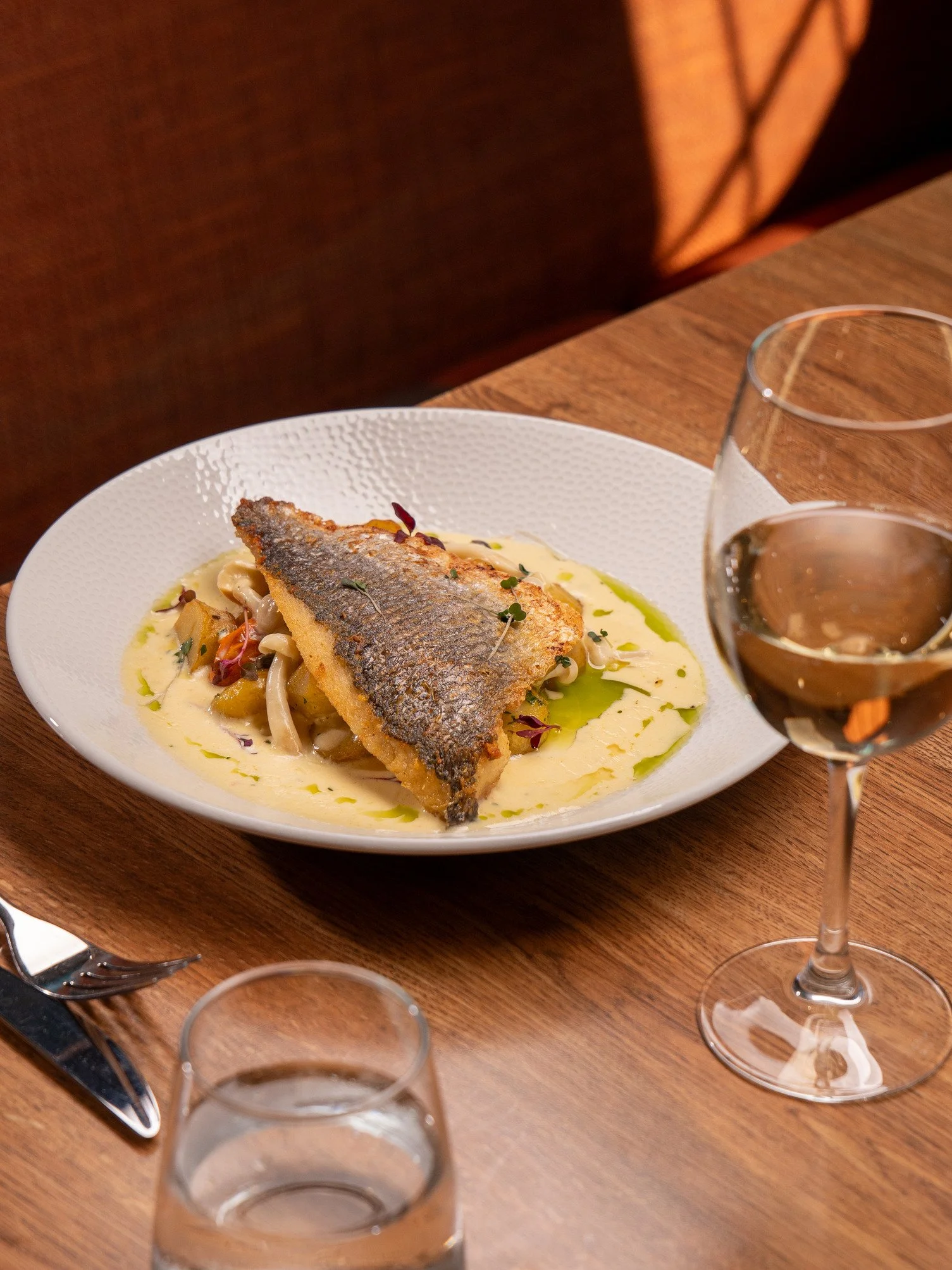 Grilled fish fillet on a bed of creamy sauce with vegetables, served on a white plate. A glass of white wine and a silverware set are nearby on a wooden table.
