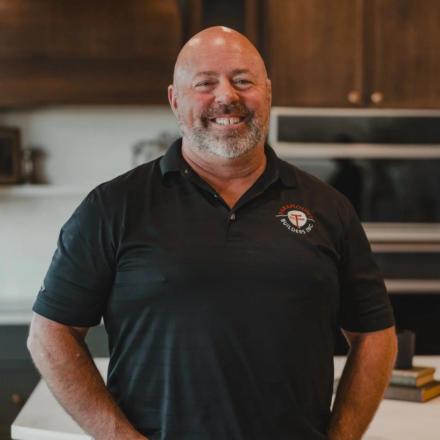 A man with a shaved head and gray beard wearing a black polo shirt with a logo that reads 'Paramount Remodeling Co., Inc.' in a kitchen with dark wood cabinets and stainless steel appliances.