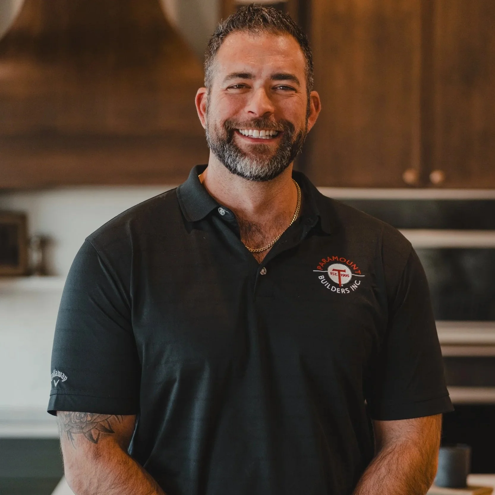 A man with dark hair and a beard, smiling with crossed arms, wearing a black polo shirt with 'Paramount Re-Modeling Co. Inc.' logo, standing in a modern kitchen.