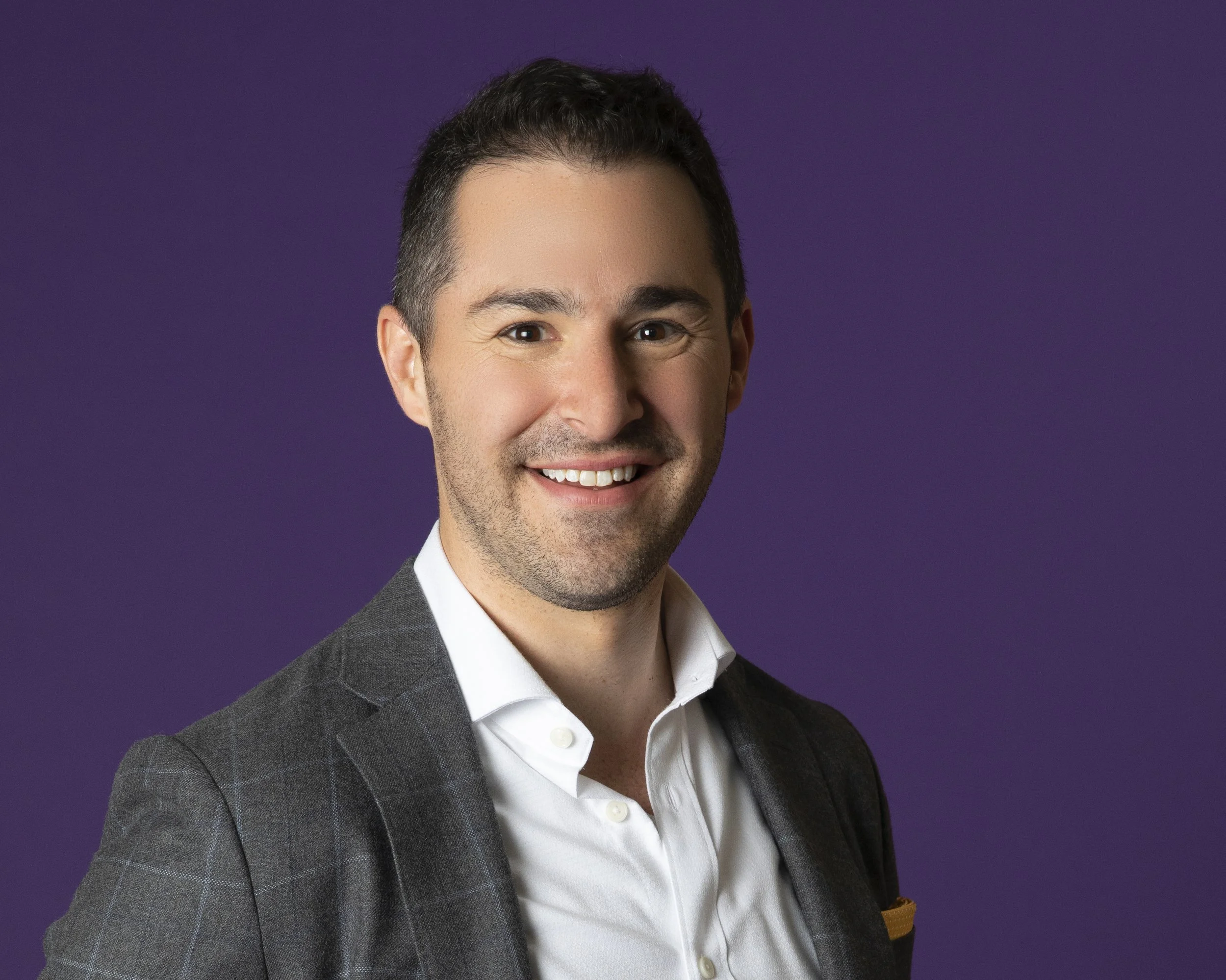 Close-up portrait of a smiling man in a suit and white shirt against a purple background.