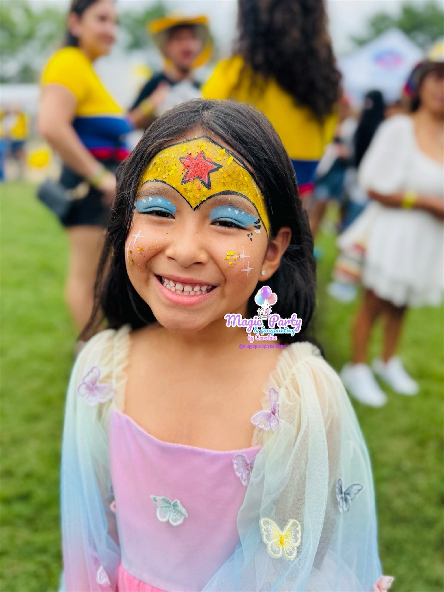 Young girl with colorful face paint and butterfly dress, smiling at an outdoor event.