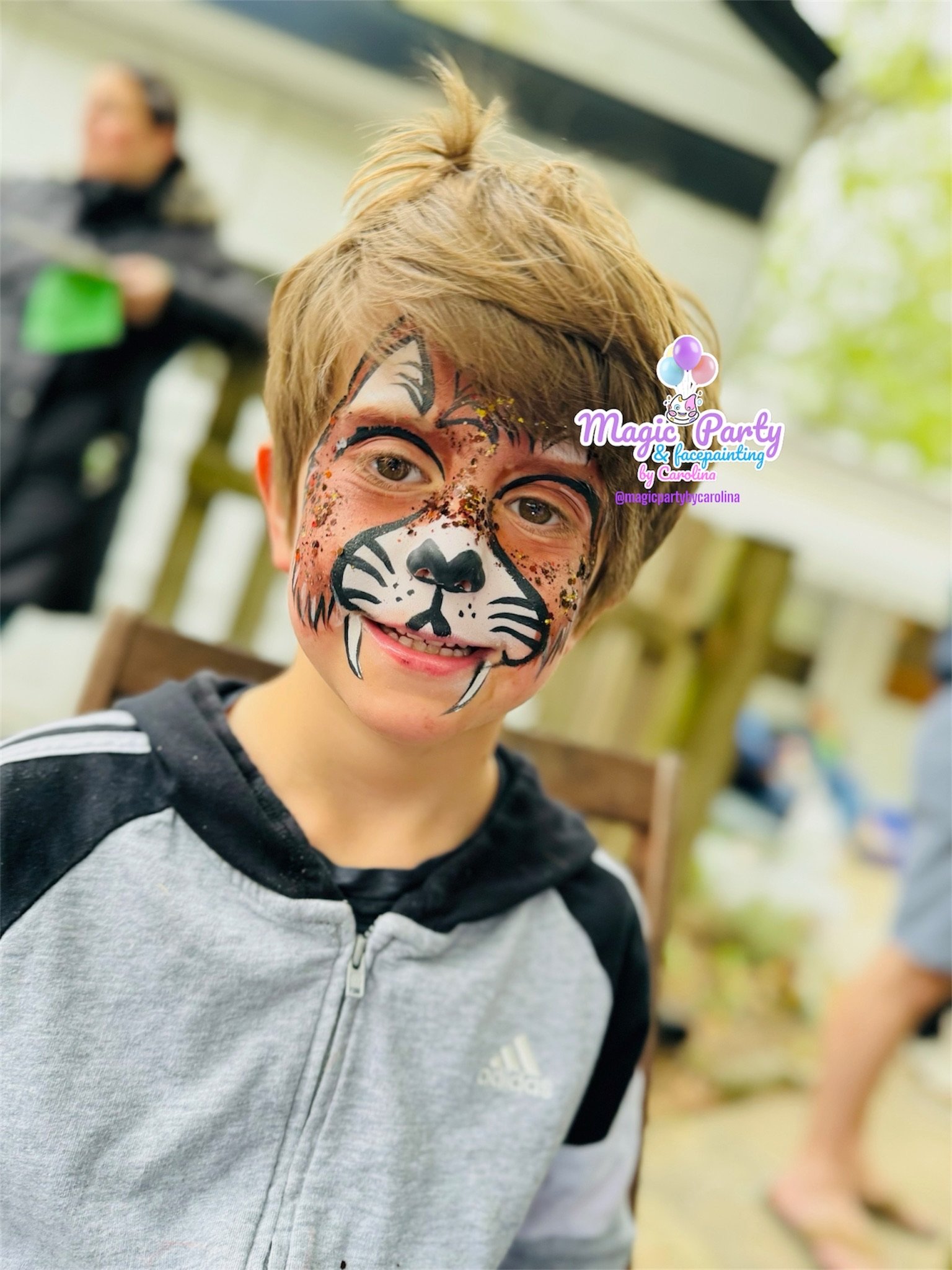 Child with tiger face paint, wearing a gray and black zip-up hoodie, smiling outdoors.