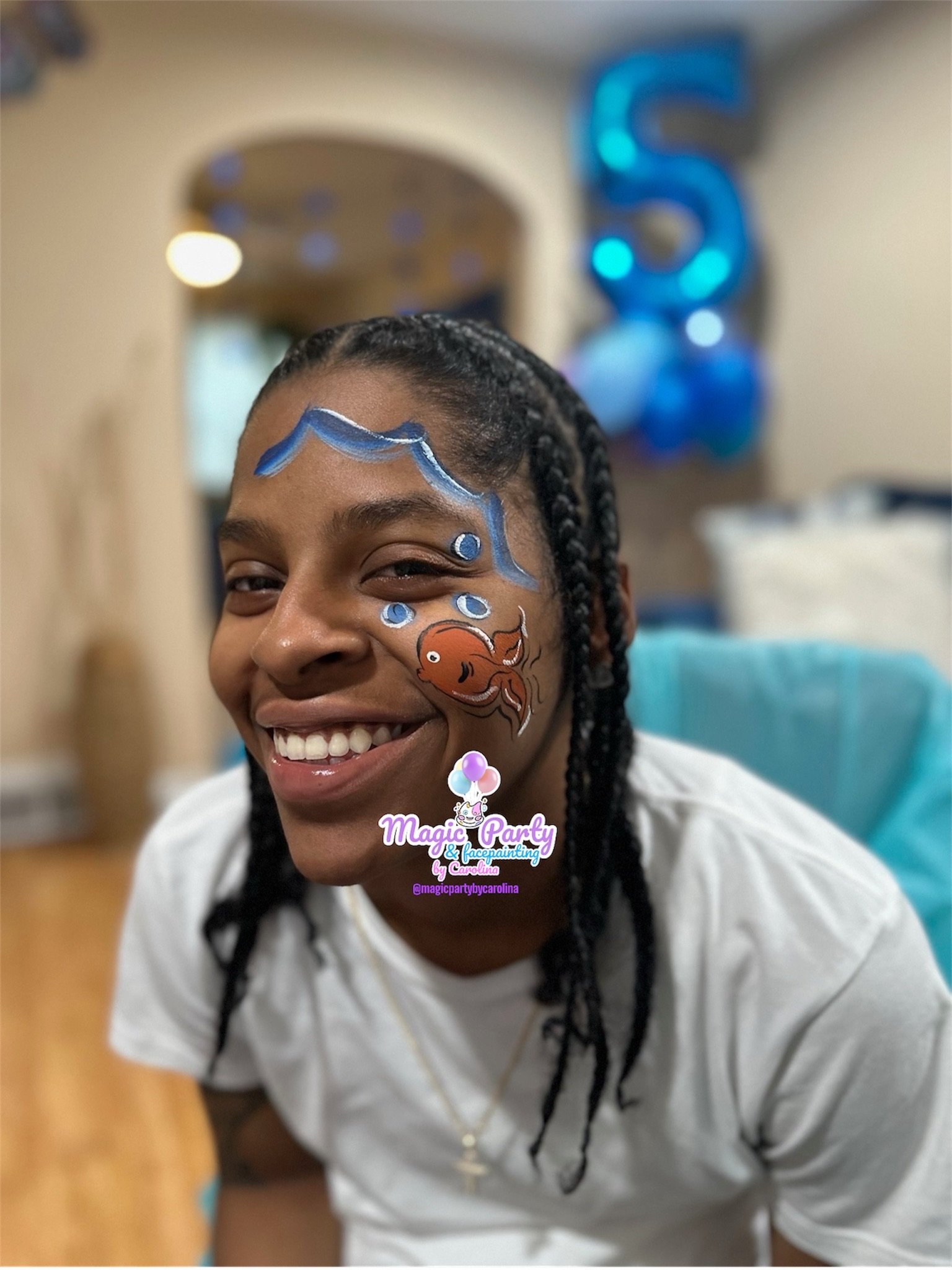 Person with face paint featuring a fish and waves, smiling indoors. Blue number 5 balloons in background.