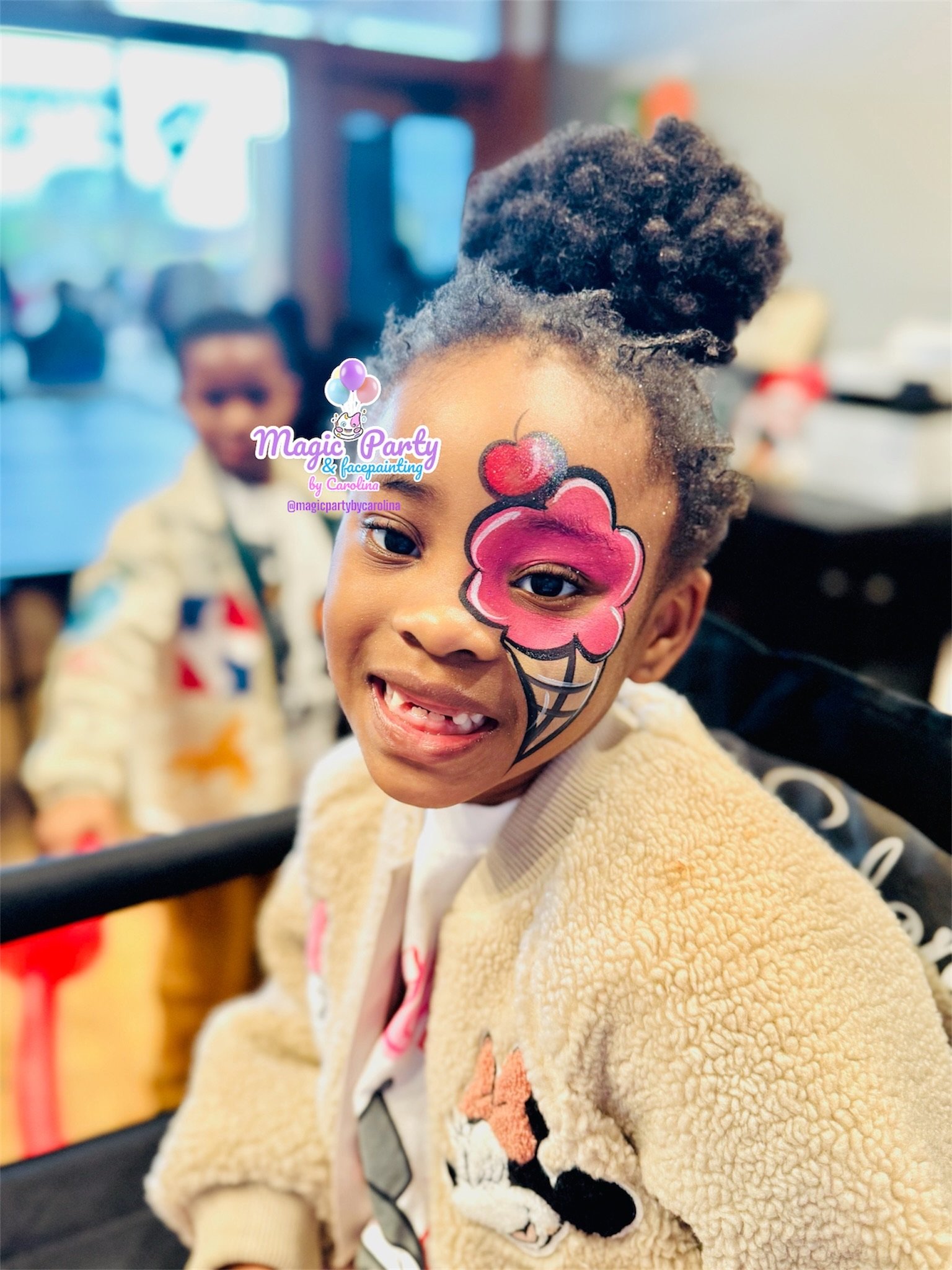 Smiling child with a pink face paint design, wearing a beige fleece jacket, in an indoor setting with another child blurred in the background.