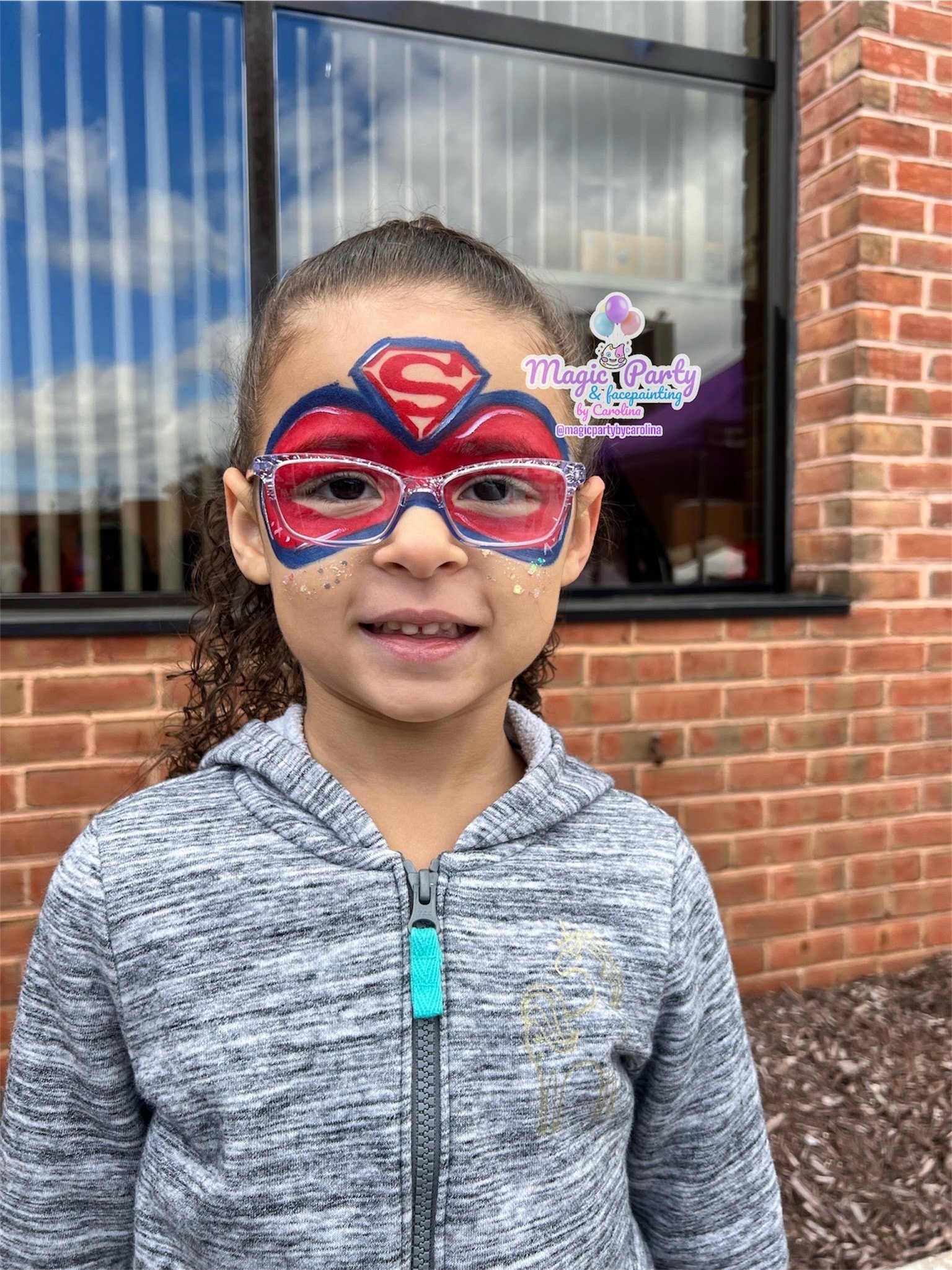 Child with Superman face paint wearing glasses, standing outside near a brick wall.