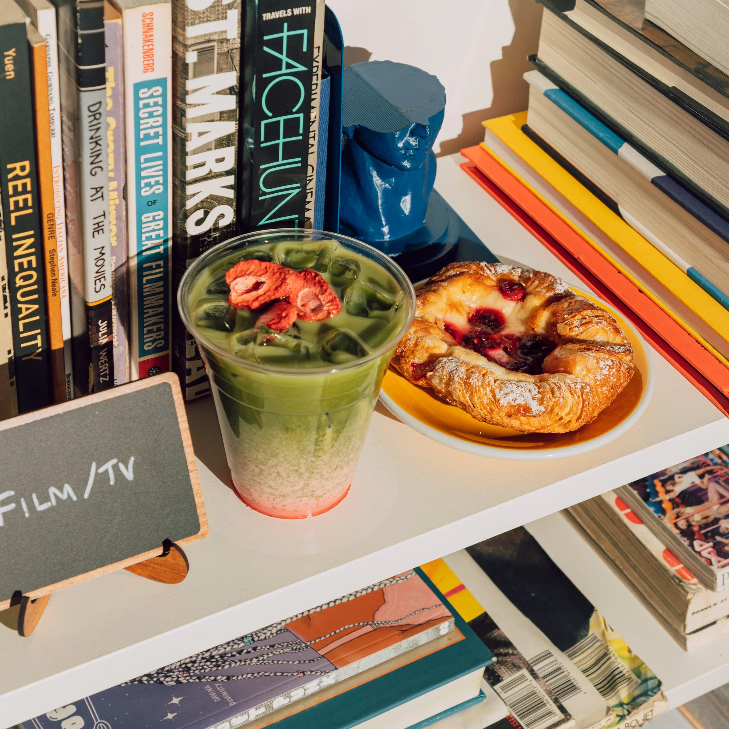 Bookshelves filled with various books, a blue sculpture of a person's head, a clear cup of iced green tea with red fruit garnish, and a plate of pastry with berries.