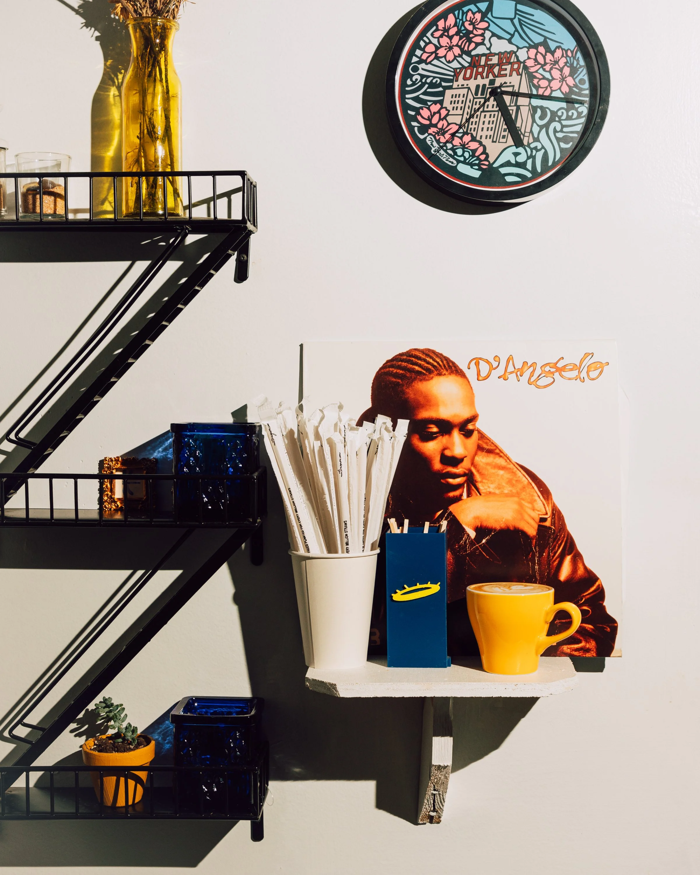Decorative wall shelf with a yellow vase with dried flowers, a small plant in an orange pot, a blue glass container, a gold chain, a blue box, a yellow mug, and a poster of a woman with braids. A round wall clock with a floral and cityscape design reads 4:12.
