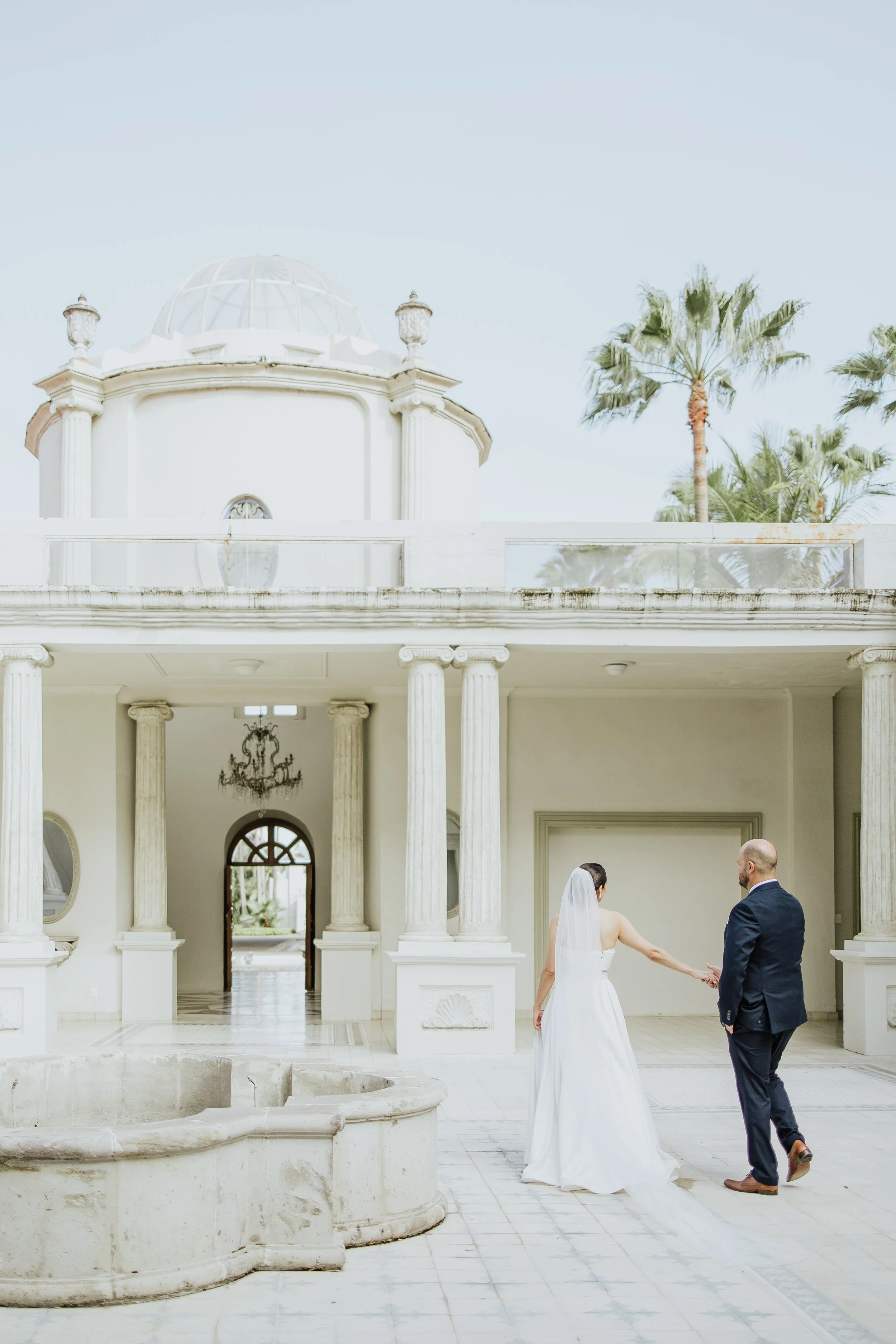 Pareja de novios en una boda, la novia con vestido blanco y velo, el novio con traje oscuro, caminando en un patio con columpios y columnas blancas, en un lugar con palmeras y arquitectura elegante.