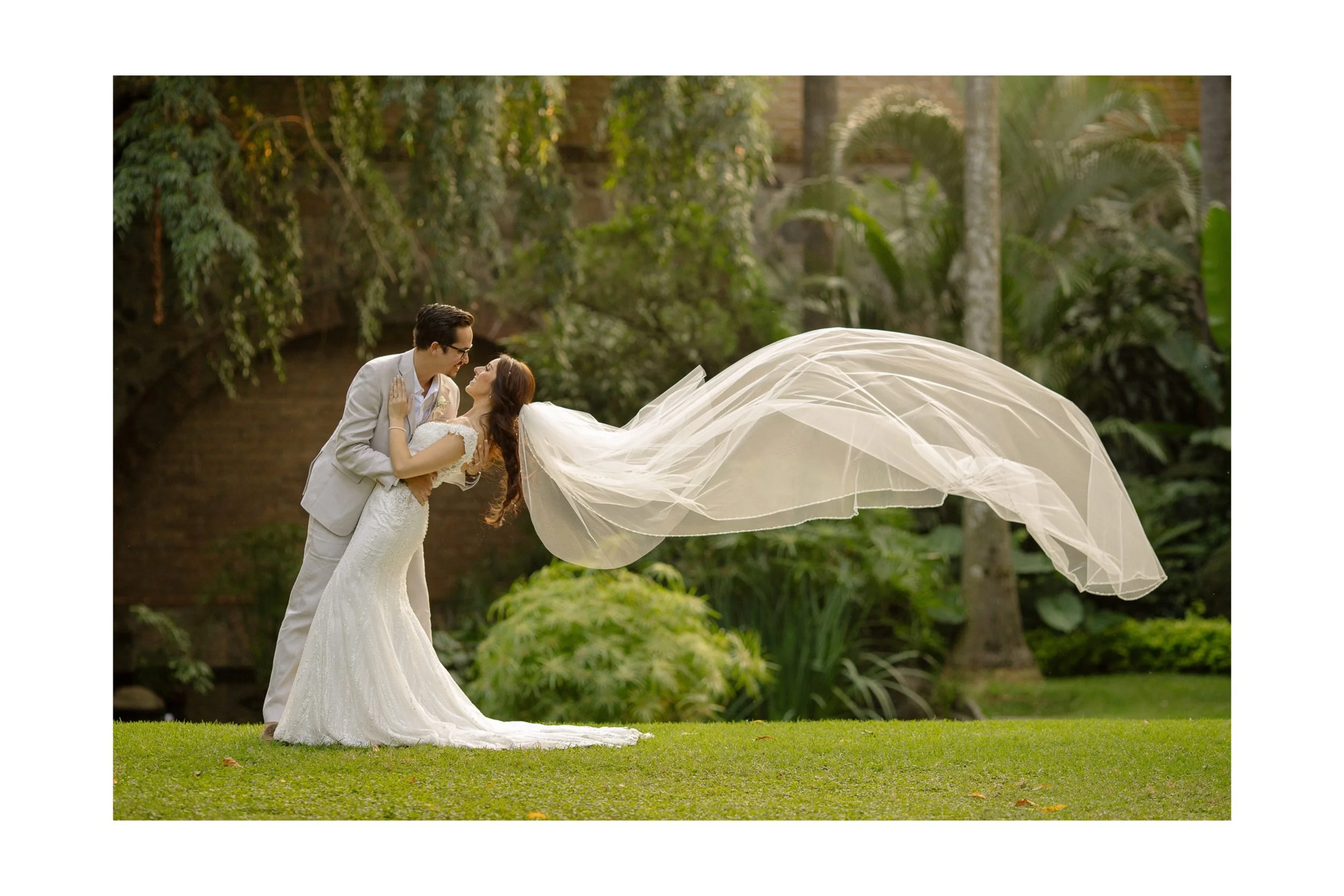 Bride and groom kissing in a garden, with bride's veil flowing in the wind.
