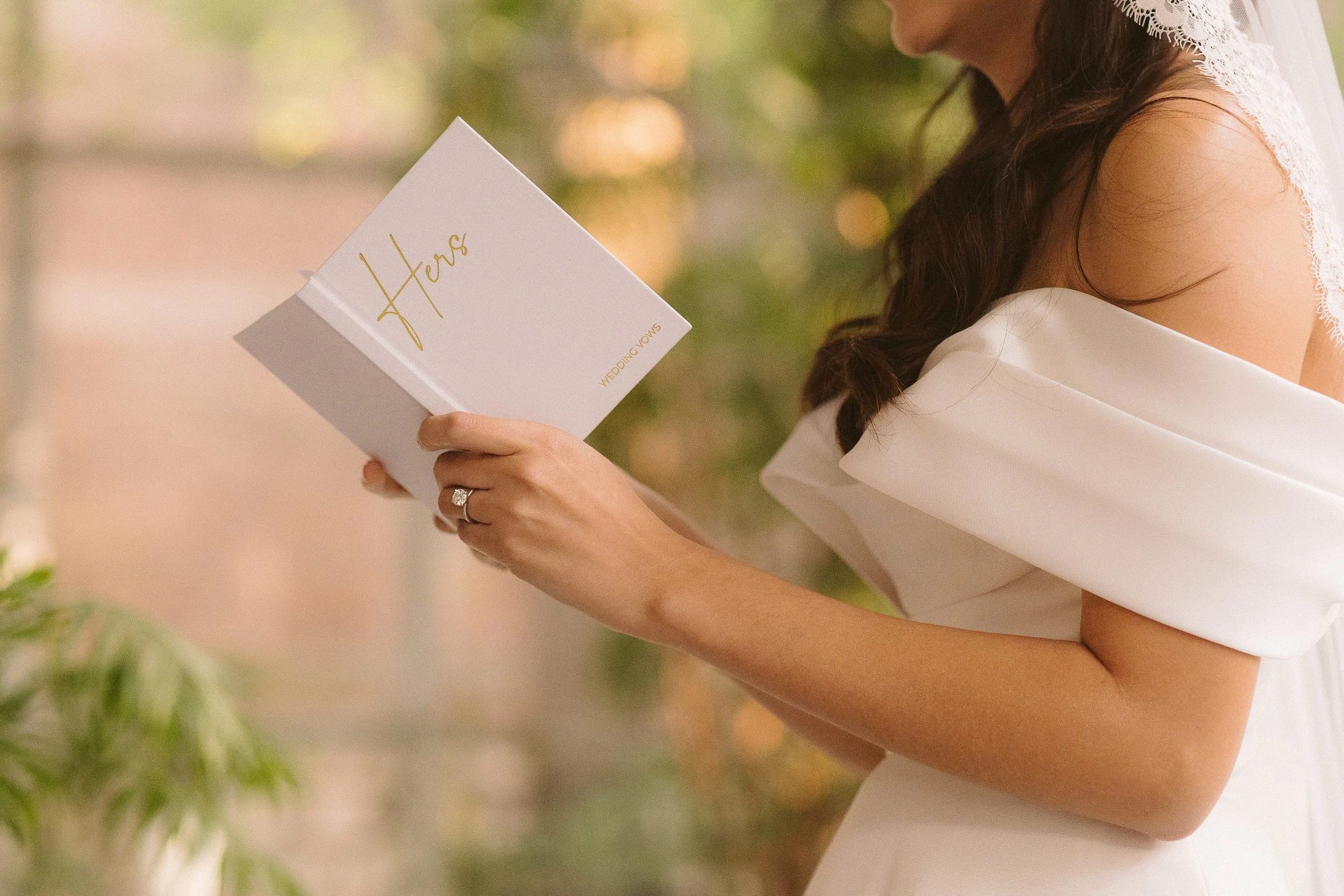 Mujer leyendo una tarjeta de bodas, vestida con un vestido blanco y un velo, en un entorno natural con luz suave y decoraciones verdes.