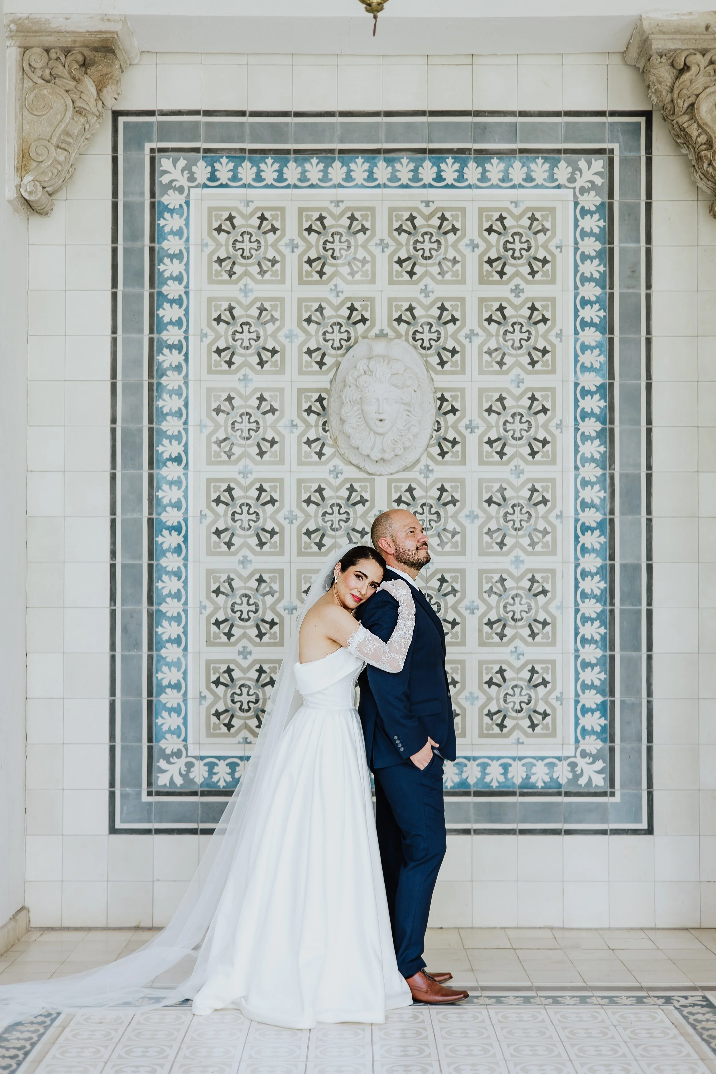 Pareja de novios en una sesión de fotos de boda, la novia abraza al novio y ambos están de perfil frente a un fondo decorativo con azulejos. Ella viste un vestido blanco y él un traje azul oscuro.