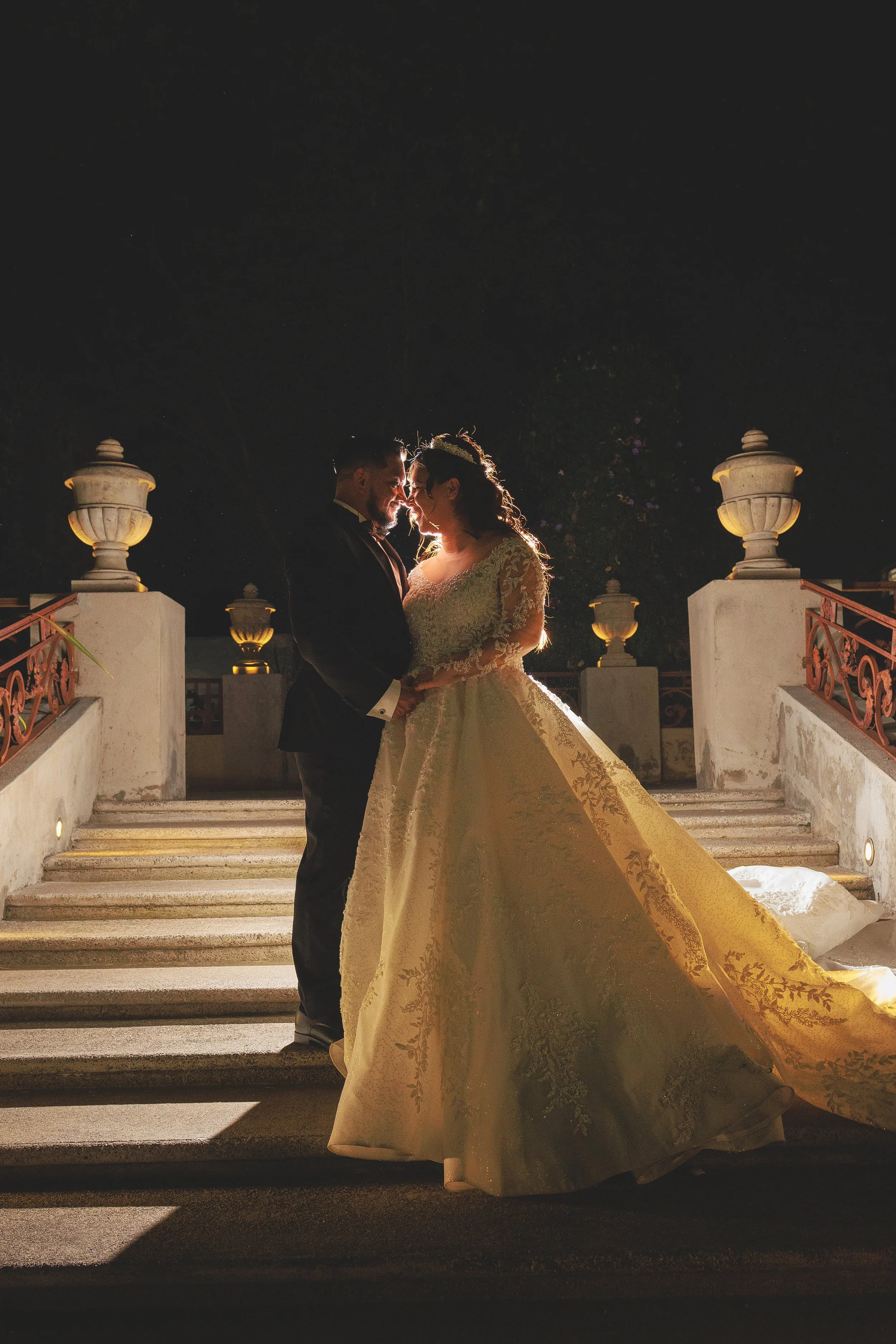 Una pareja de novios de pie en unas escaleras en la noche, con iluminación suave detrás. La novia usa un vestido de boda elegante y la pareja se mira íntimamente.