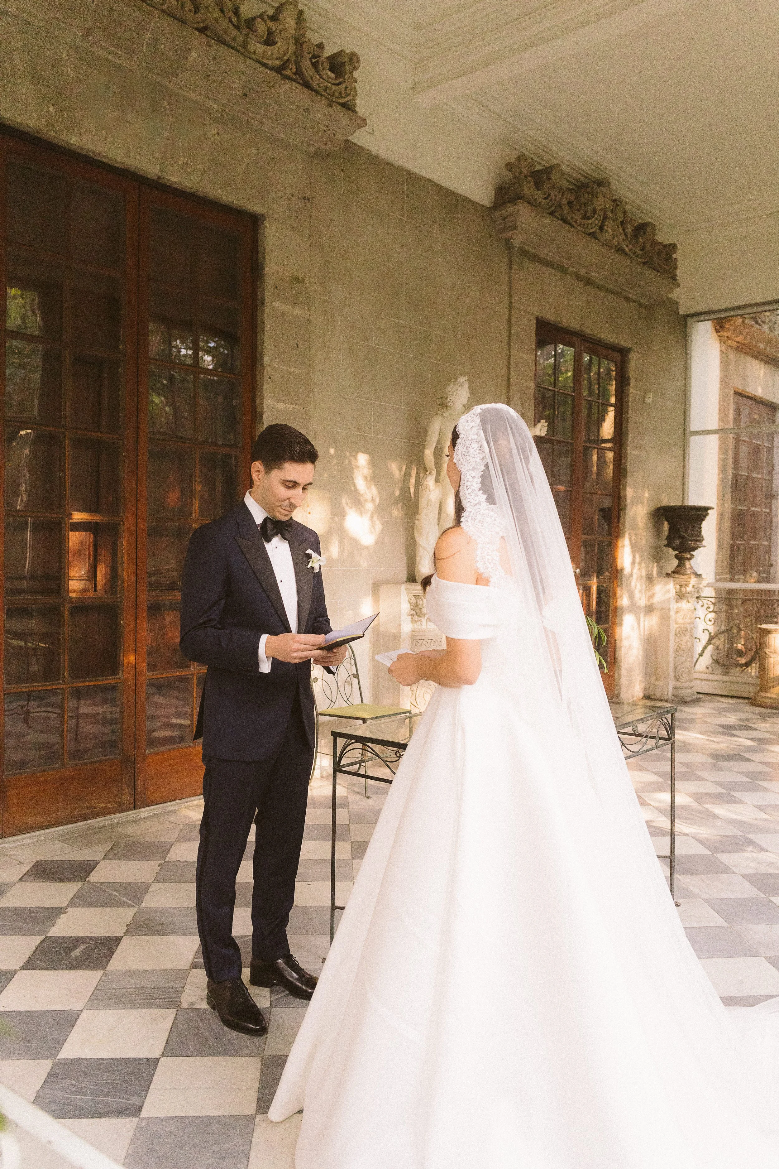Una pareja de novios realiza sus votos en una boda en un salón elegante con paredes de piedra, ventanas de madera y decoraciones clásicas, como estatuas y jarrones grandes.