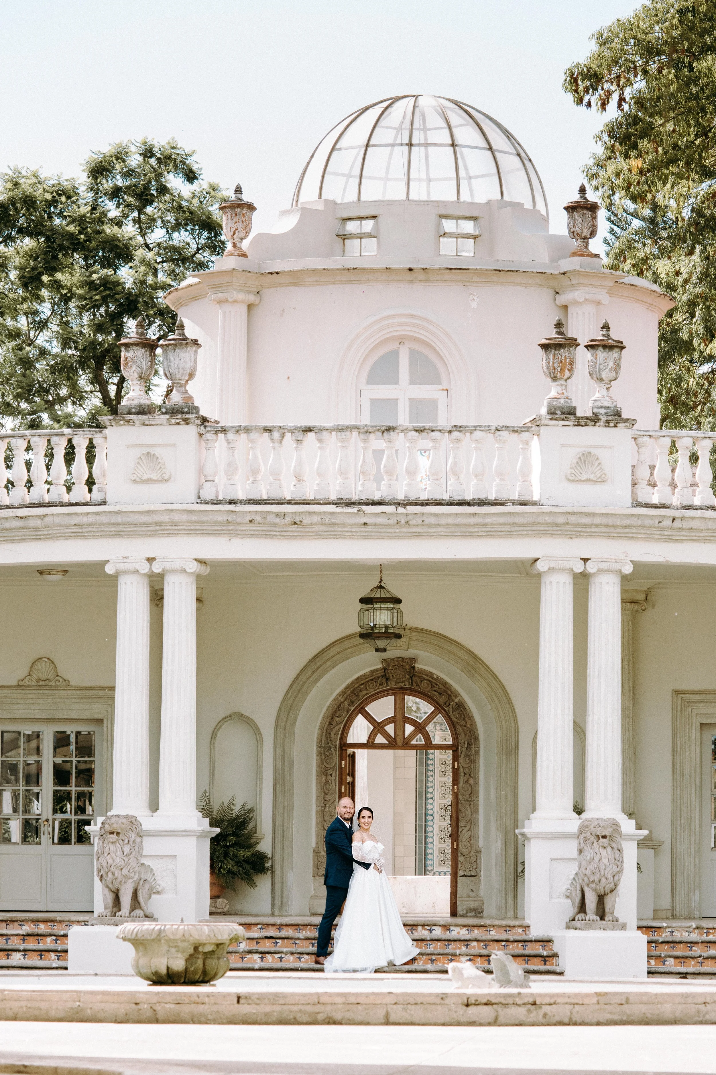 Matrimonio en una mansión elegante con columnas y esculturas de leones, pareja en el umbral de la entrada, ambiente soleado y árboles al fondo.