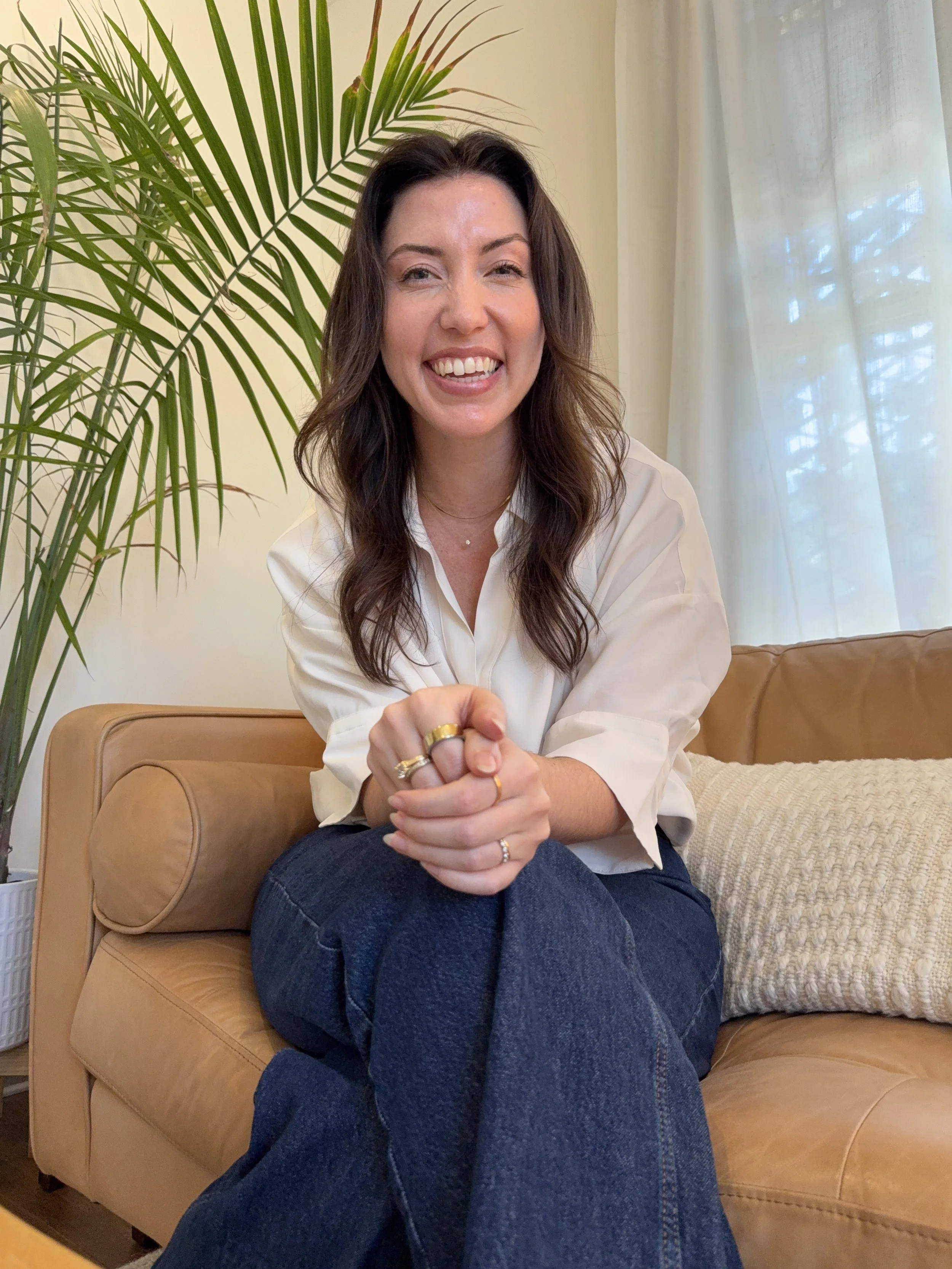 A woman with long brown hair, wearing a white blouse and dark blue jeans, sitting on a tan leather couch in a living room, smiling and looking at the camera. There is a large green plant to her left and a window with light-colored curtains behind her.