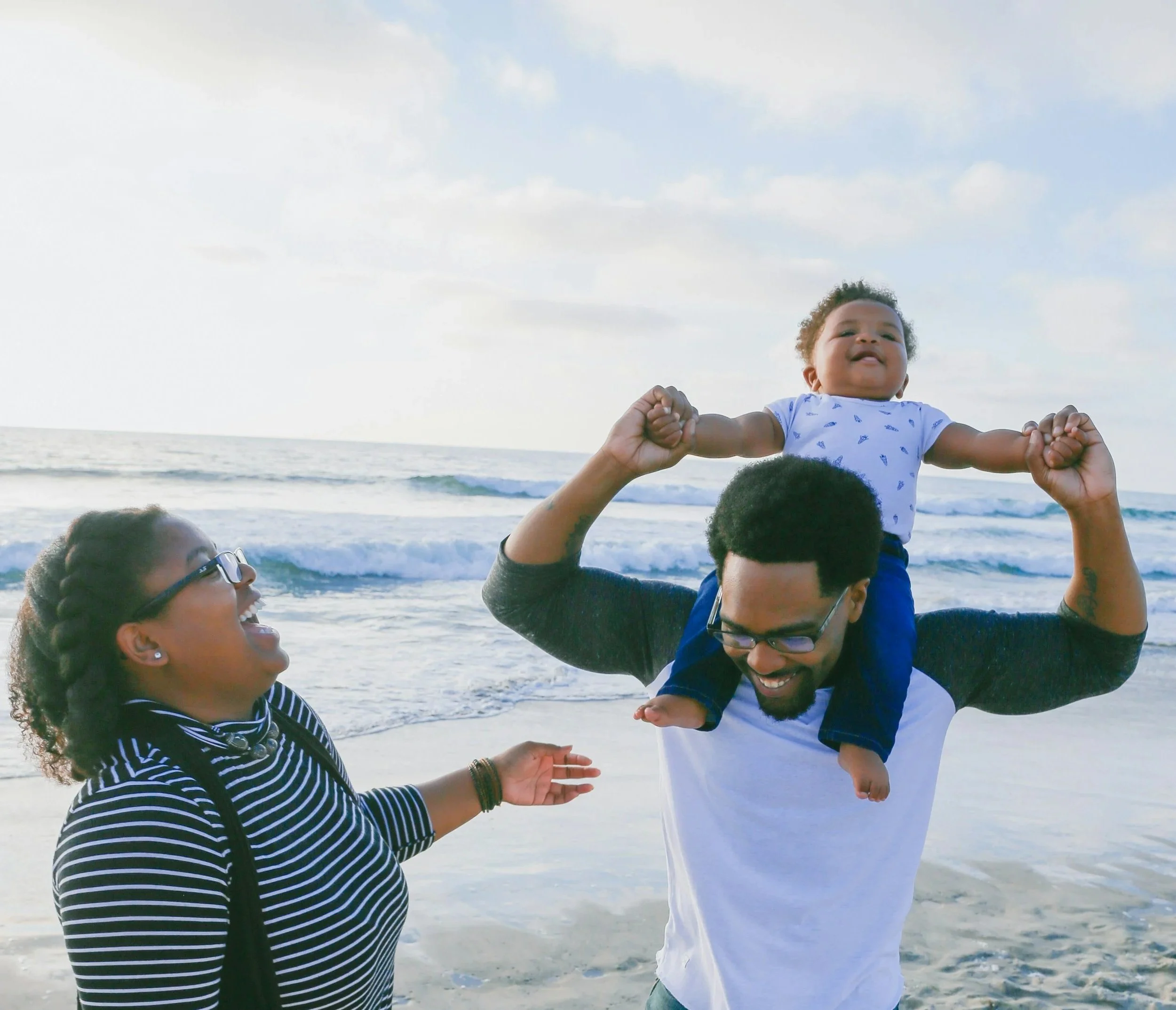A group of adults holding a smiling child in the air at the beach, with waves and a cloudy sky in the background.