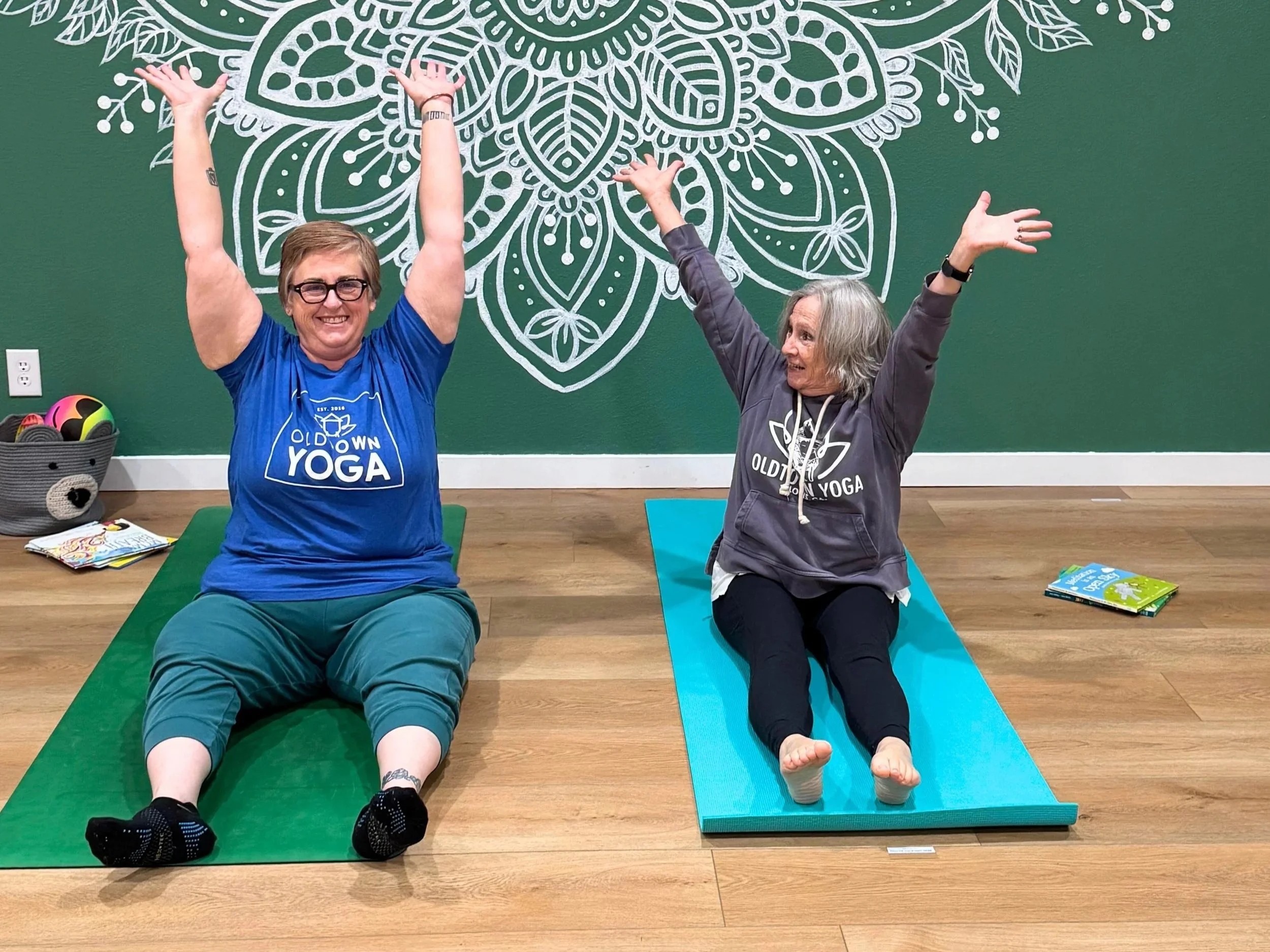 Two women doing yoga on mats in a studio, smiling with their arms raised. One in a blue shirt, the other in a gray hoodie, with a green wall and white decorative design behind them.