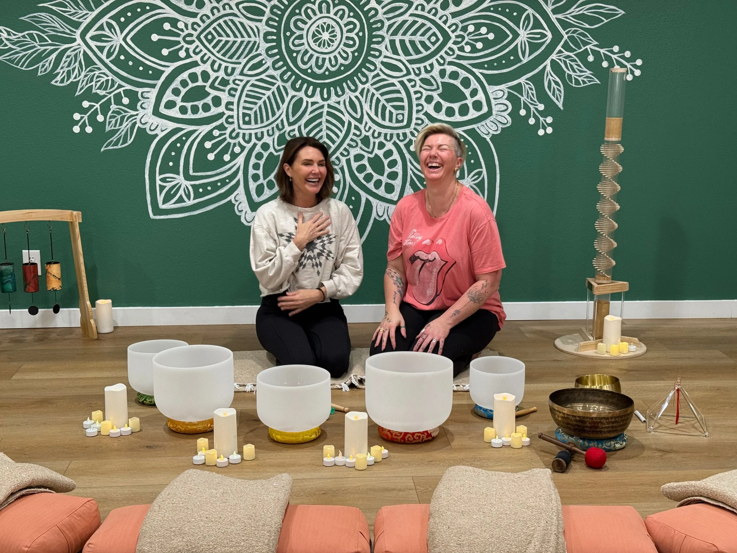 Two women sitting on the floor, laughing, surrounded by singing bowls, candles, and meditation tools, with a green wall and white decorative mandala art behind them.