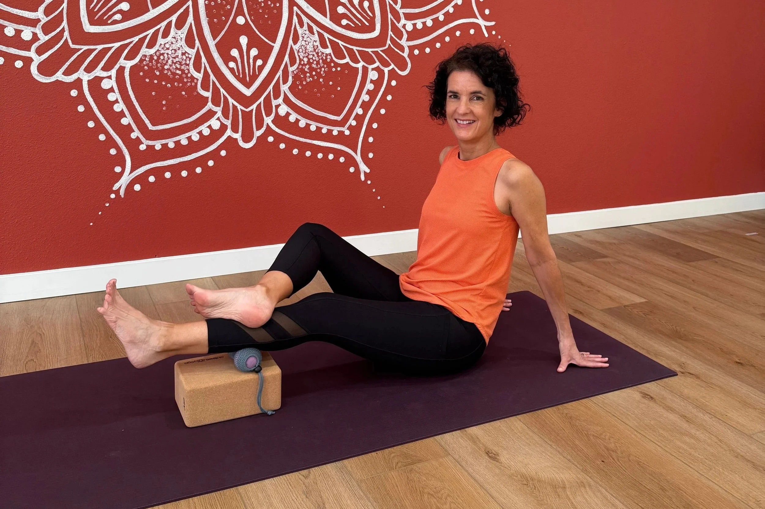 A woman in an orange sleeveless top and black leggings is seated on a yoga mat in a fitness studio. She has one leg extended onto a foam roller, with her foot resting on a block, and is smiling at the camera. The studio has a red wall with white decorative chalk art.