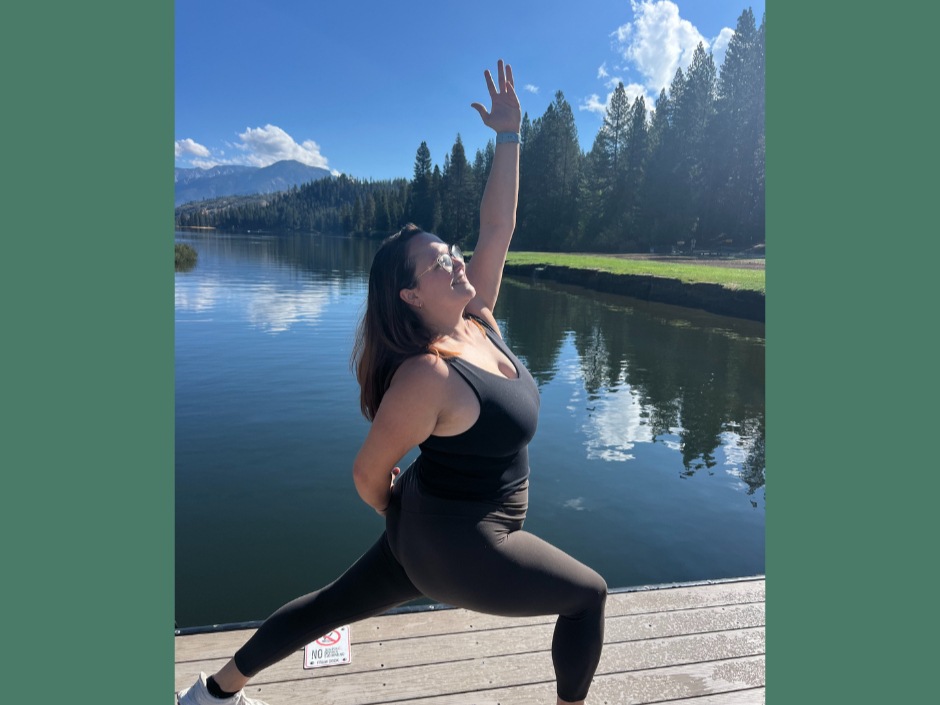 A woman in activewear doing a yoga pose on a dock by a lake, with trees and mountains in the background under a partly cloudy sky.