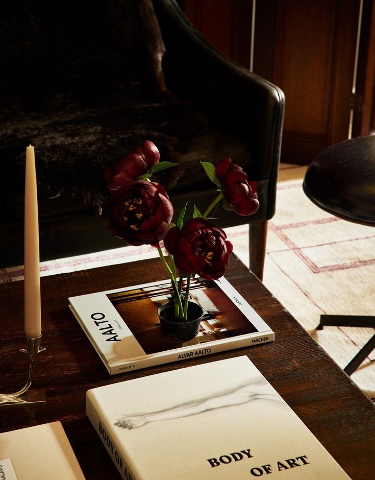 A dark wood table with a white candle in a silver holder, a black vase with burgundy tulips, and two books titled 'Alvar Aalto' and 'Body of Art.'