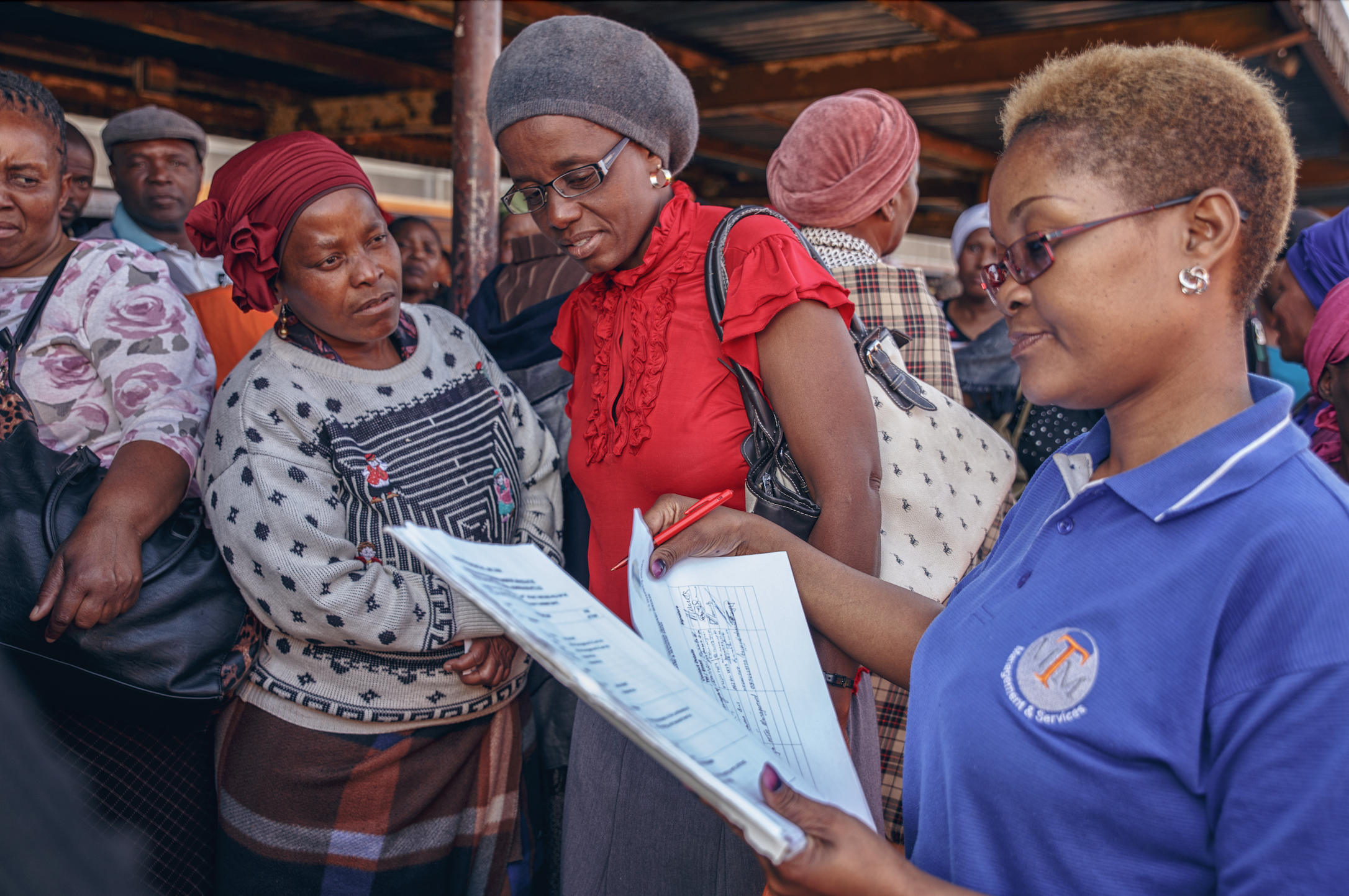 A woman in a blue shirt with a logo on it is holding a clipboard and writing, while a group of women look on at an outdoor market or event.