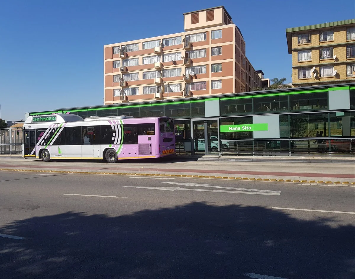 A modern bus parked outside a building with a glass facade at a bus stop labeled Nana Sita. The bus has a white, purple, and green color scheme. There are residential buildings in the background under a clear, blue sky.