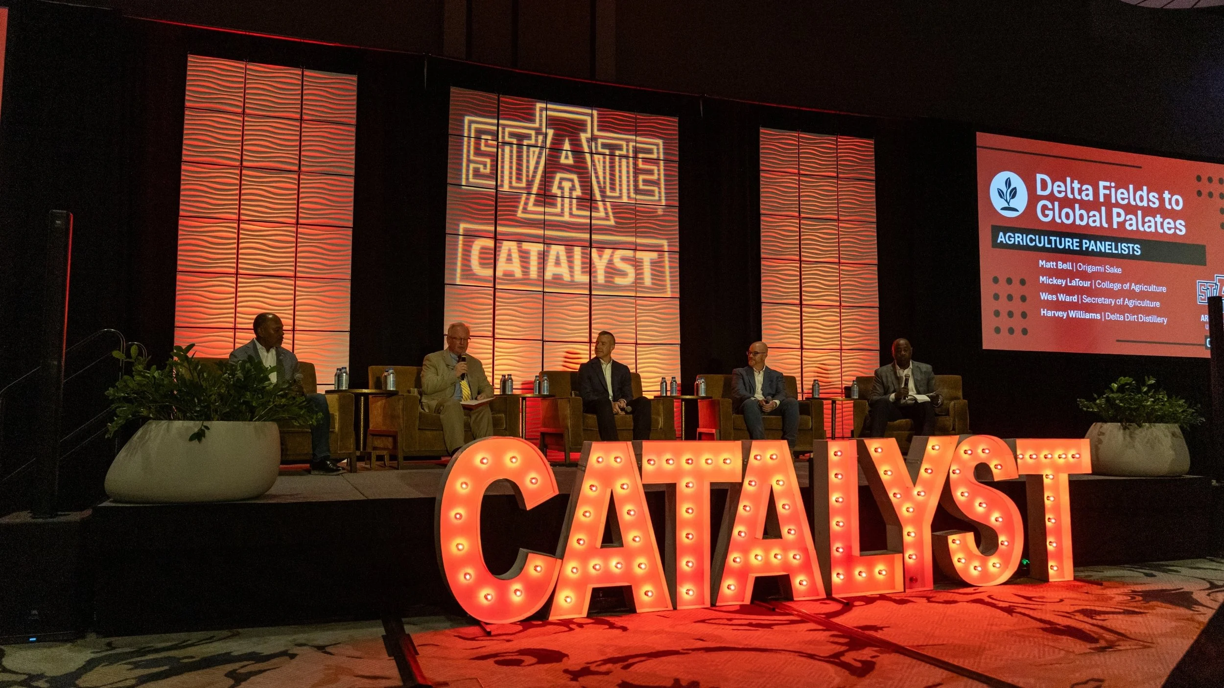 A panel discussion at a conference with five men seated on stage, behind them a large screen displaying panelist names and the text 'Delta Fields to Global Palates,' and a prominent illuminated sign spelling 'CATALYST' in front of the stage.