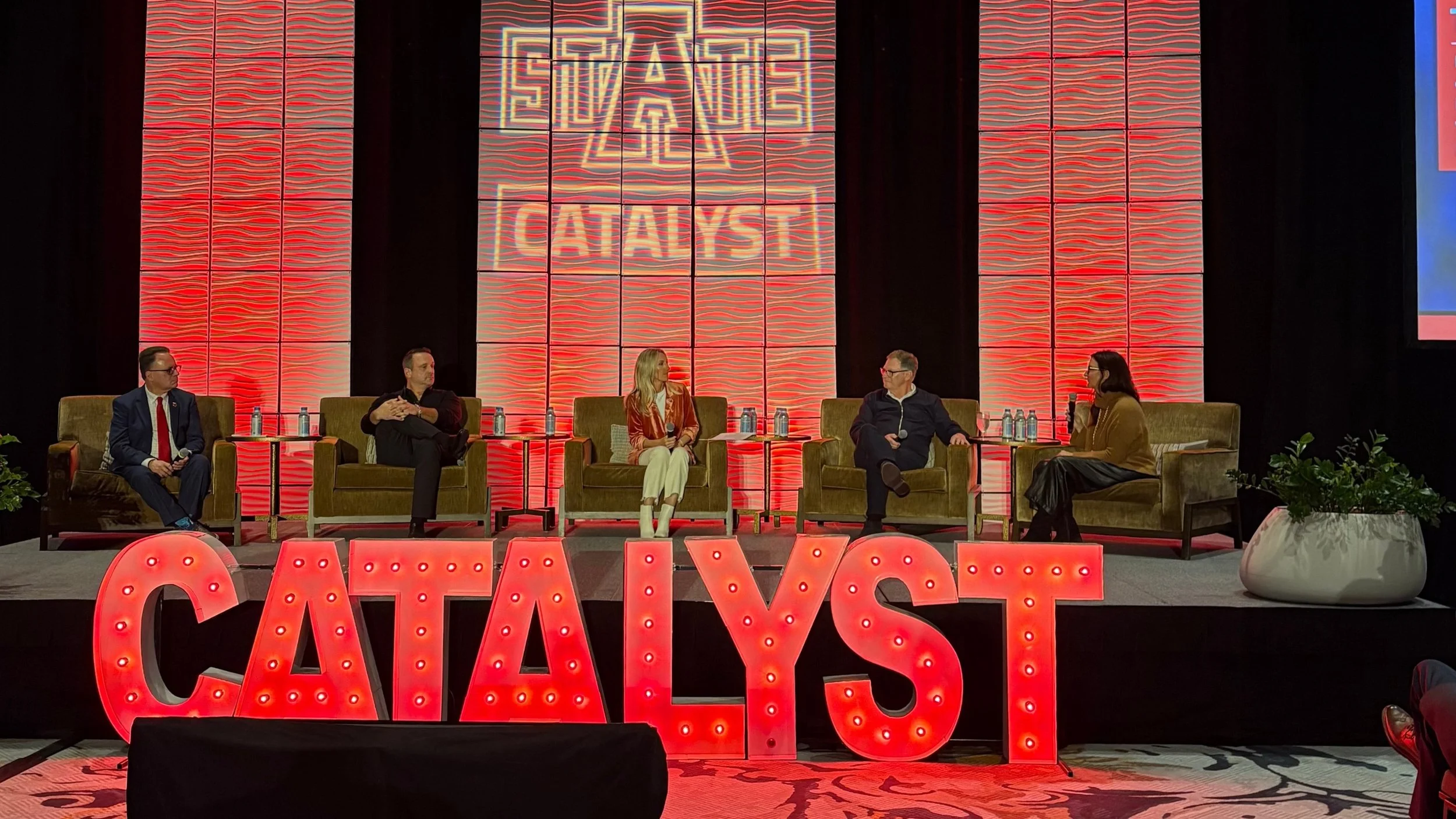 A panel discussion on stage with five people seated in armchairs, behind a large illuminated sign that reads 'CATAST' and a backdrop with the logo 'FII ATE' and the word 'CATAST.' There are small tables with water bottles beside each panelist and a p