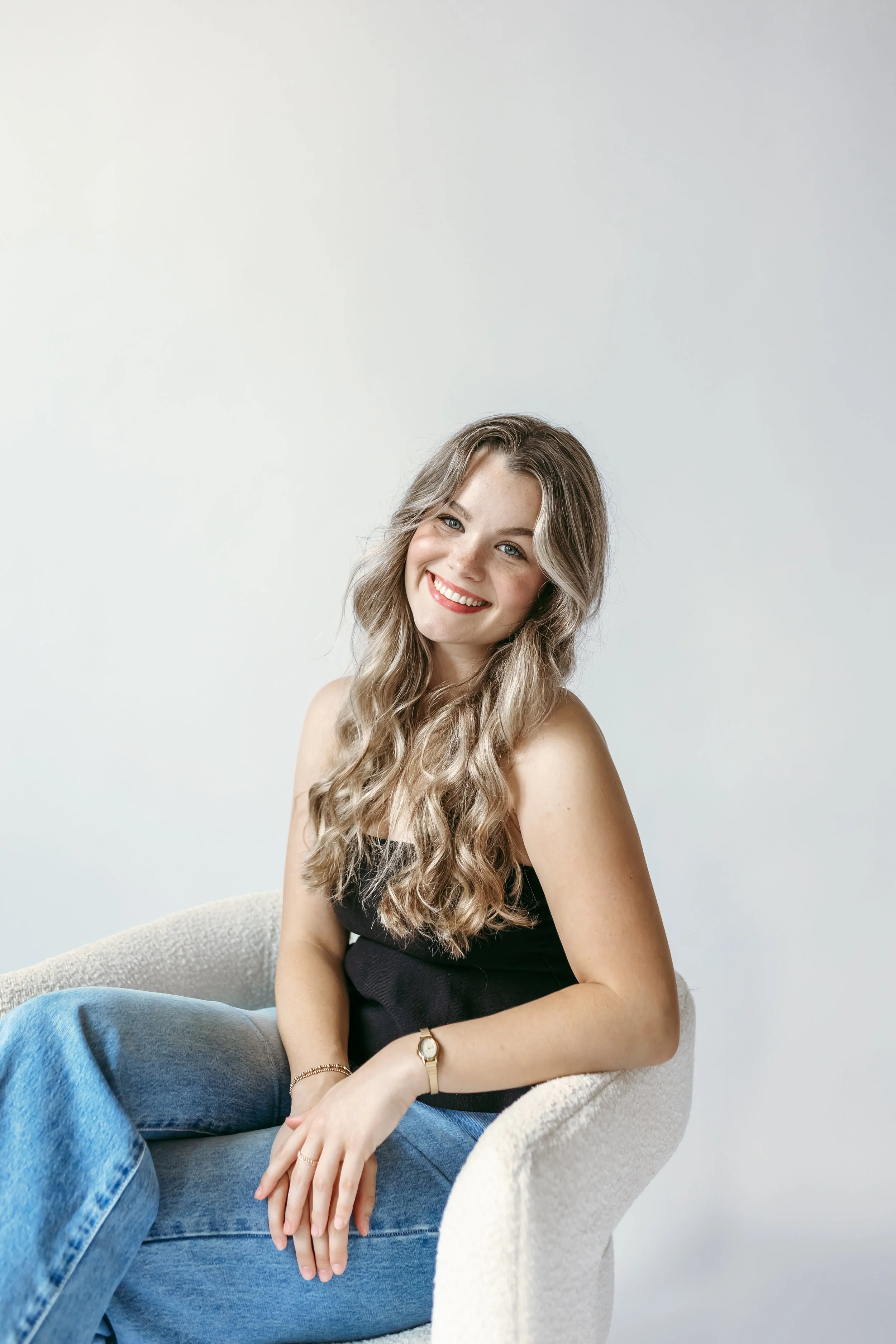 A young woman with long wavy blonde hair, smiling, sitting on a light-colored chair against a plain white wall, wearing a black tank top and blue jeans, with a watch and bracelet on her left wrist.