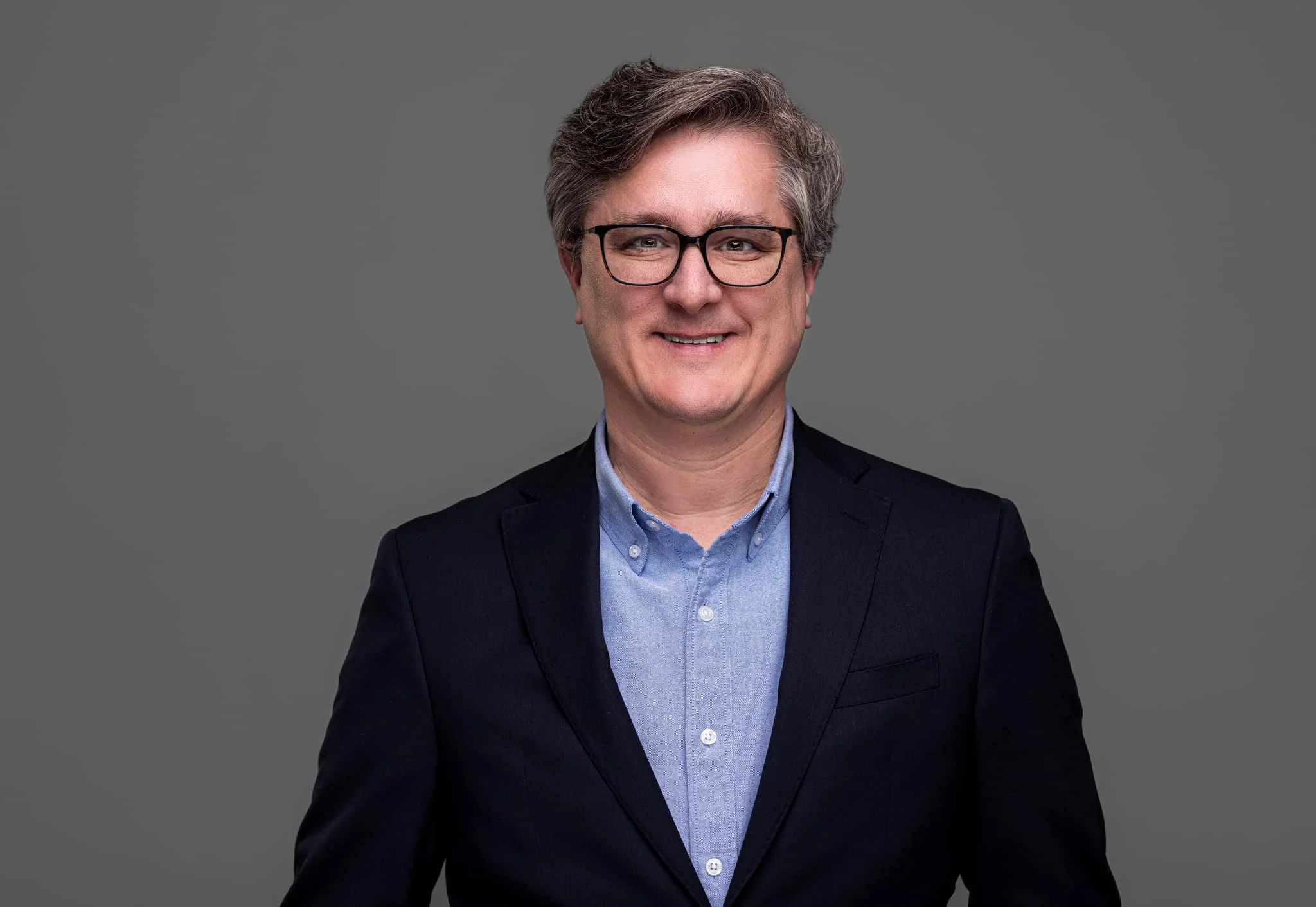 A middle-aged man with gray hair, glasses, wearing a dark blazer over a light blue button-up shirt, smiling against a gray background.