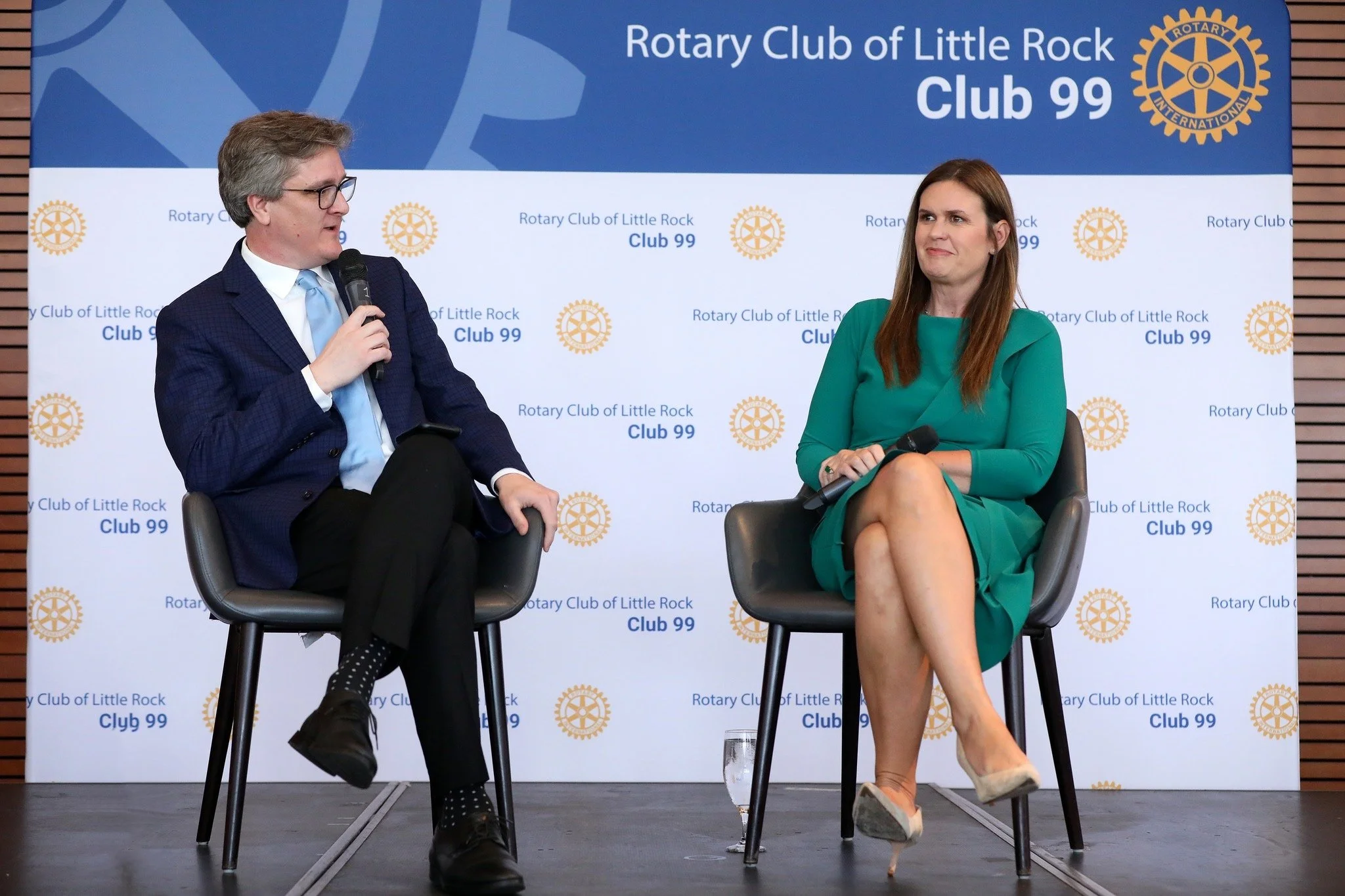 A man and woman seated on stage, engaged in conversation, at a Rotary Club event. The backdrop displays the Rotary Club of Little Rock logo and the text 'Club 99'.