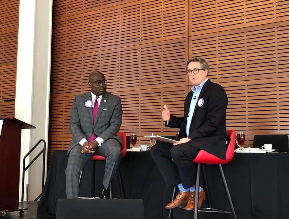 Two men in suits sitting on stage during a panel discussion at a conference or event, with a wooden-paneled wall in the background, and a table with drinks and cups behind them.