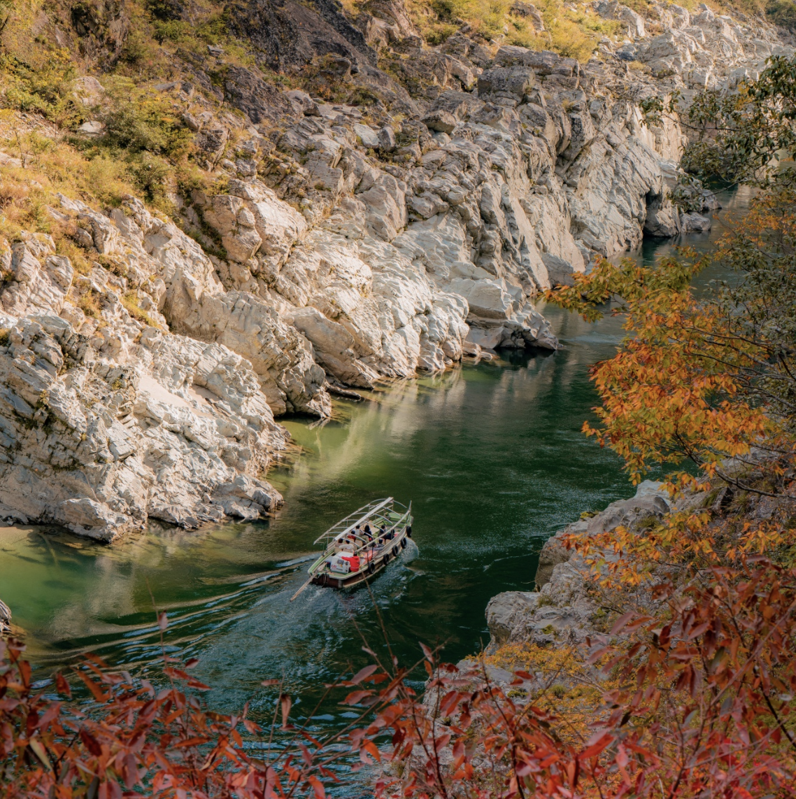 A boat with passengers navigating through a narrow river surrounded by steep rocky cliffs and autumn-colored foliage.