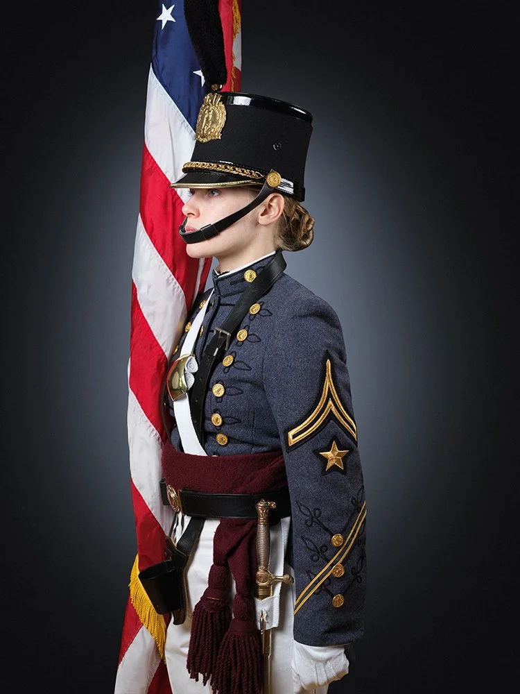 A woman dressed in a World War II army uniform holding an American flag. She is wearing a decorated uniform, a black shako hat with a gold emblem and black plume, and a headset microphone.