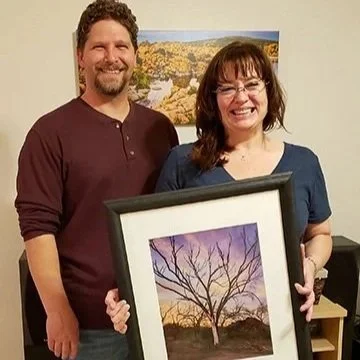 A man and woman smiling and holding a framed tree painting in an art gallery.