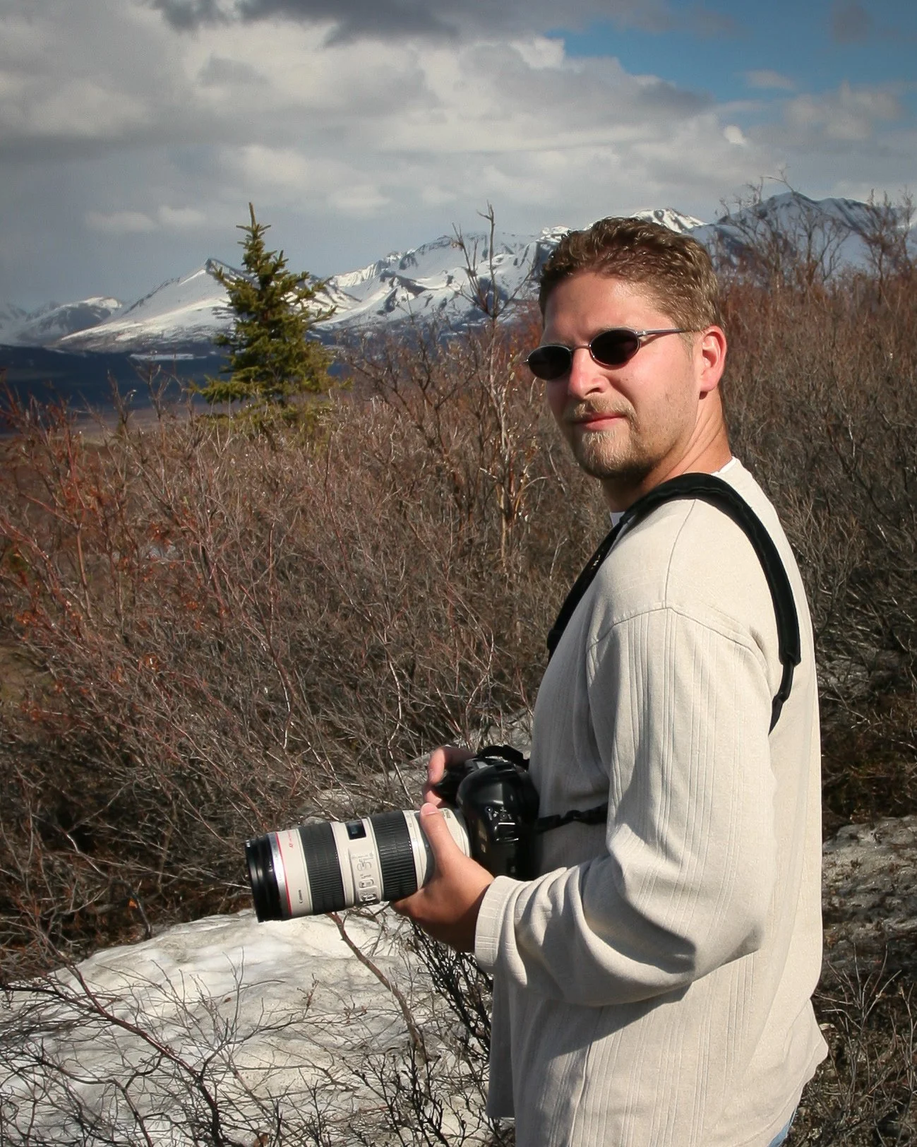 A man with sunglasses holding a professional camera with a telephoto lens outdoors in a mountainous area with snow-capped peaks, leafless bushes, and cloudy sky.