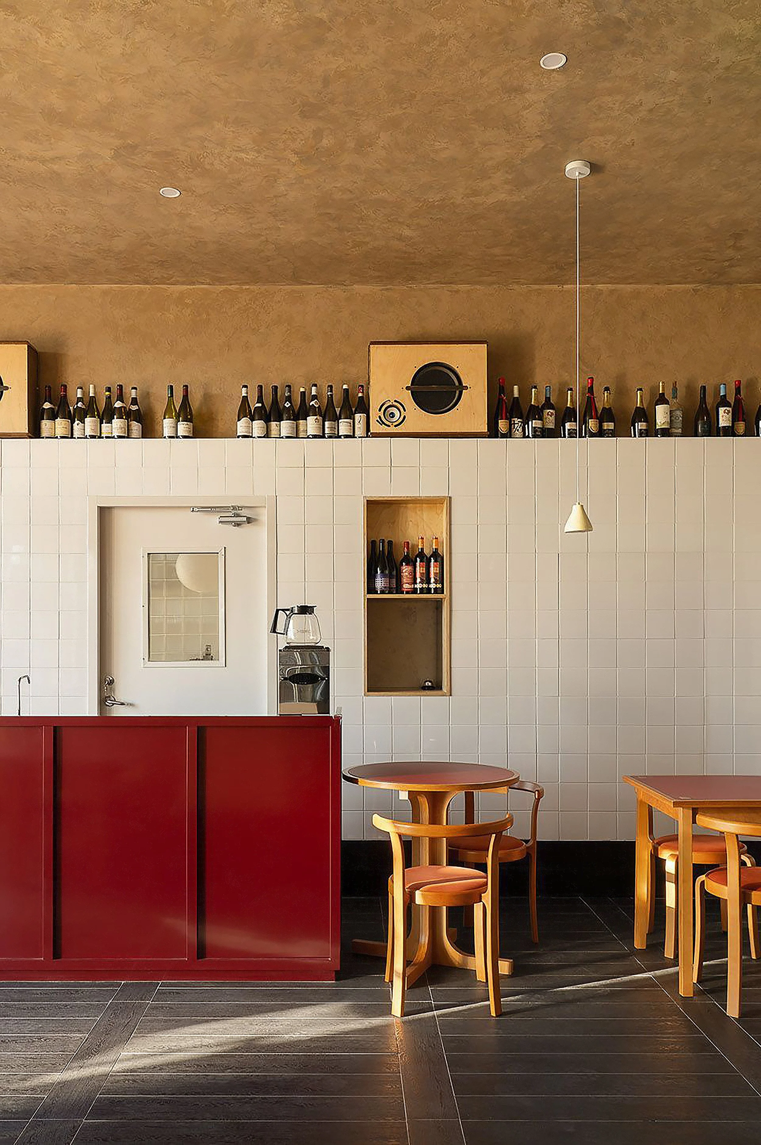 Interior of a modern cafe or restaurant with wine bottles on top of white tiled walls, wooden tables and chairs, a coffee machine, and a red partition.