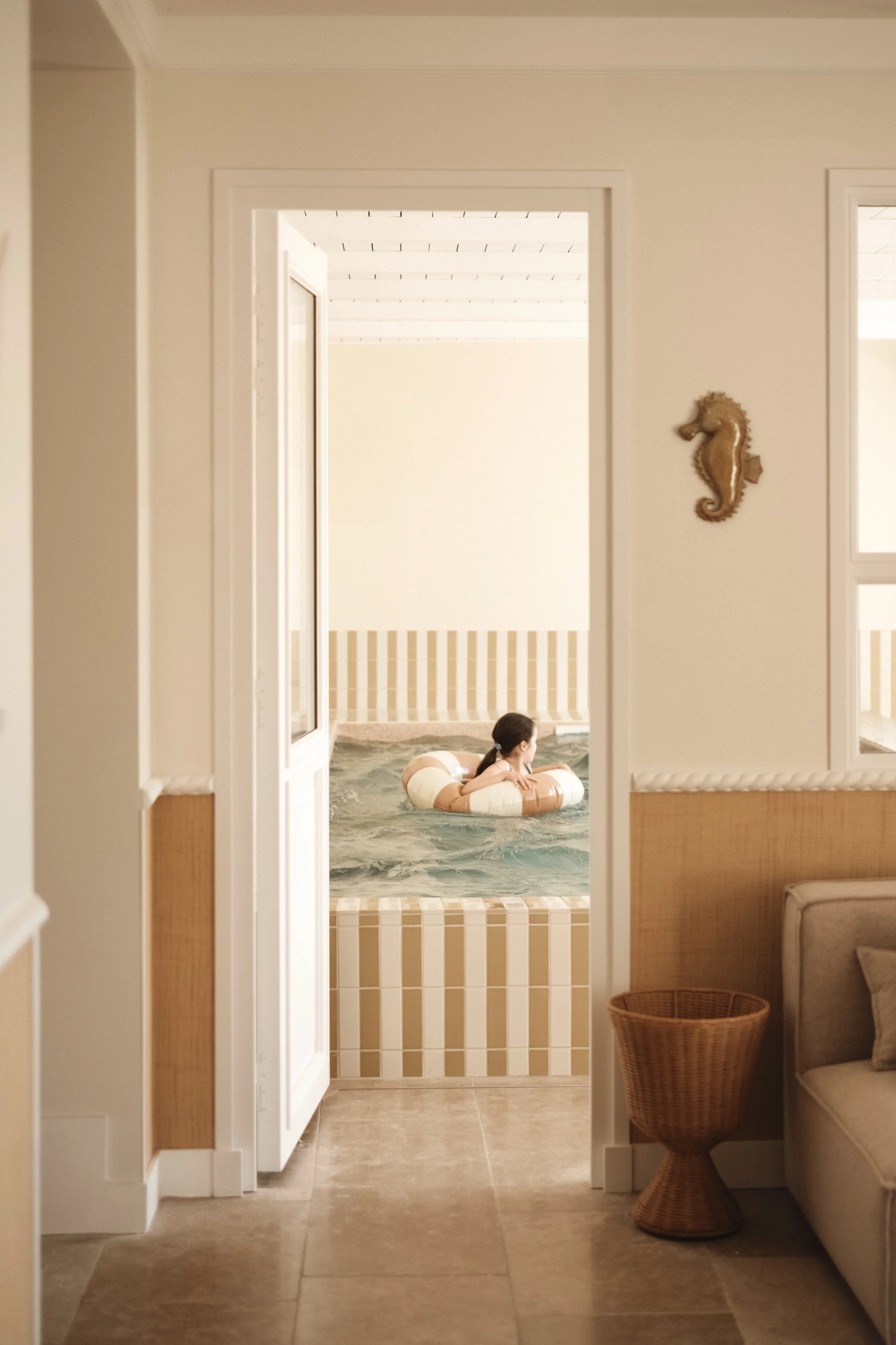 Une fille nage dans une piscine intérieure, vue depuis une pièce avec un canapé beige et une table en rotin, décorée avec une tête de hippocampe en bois sur le mur.