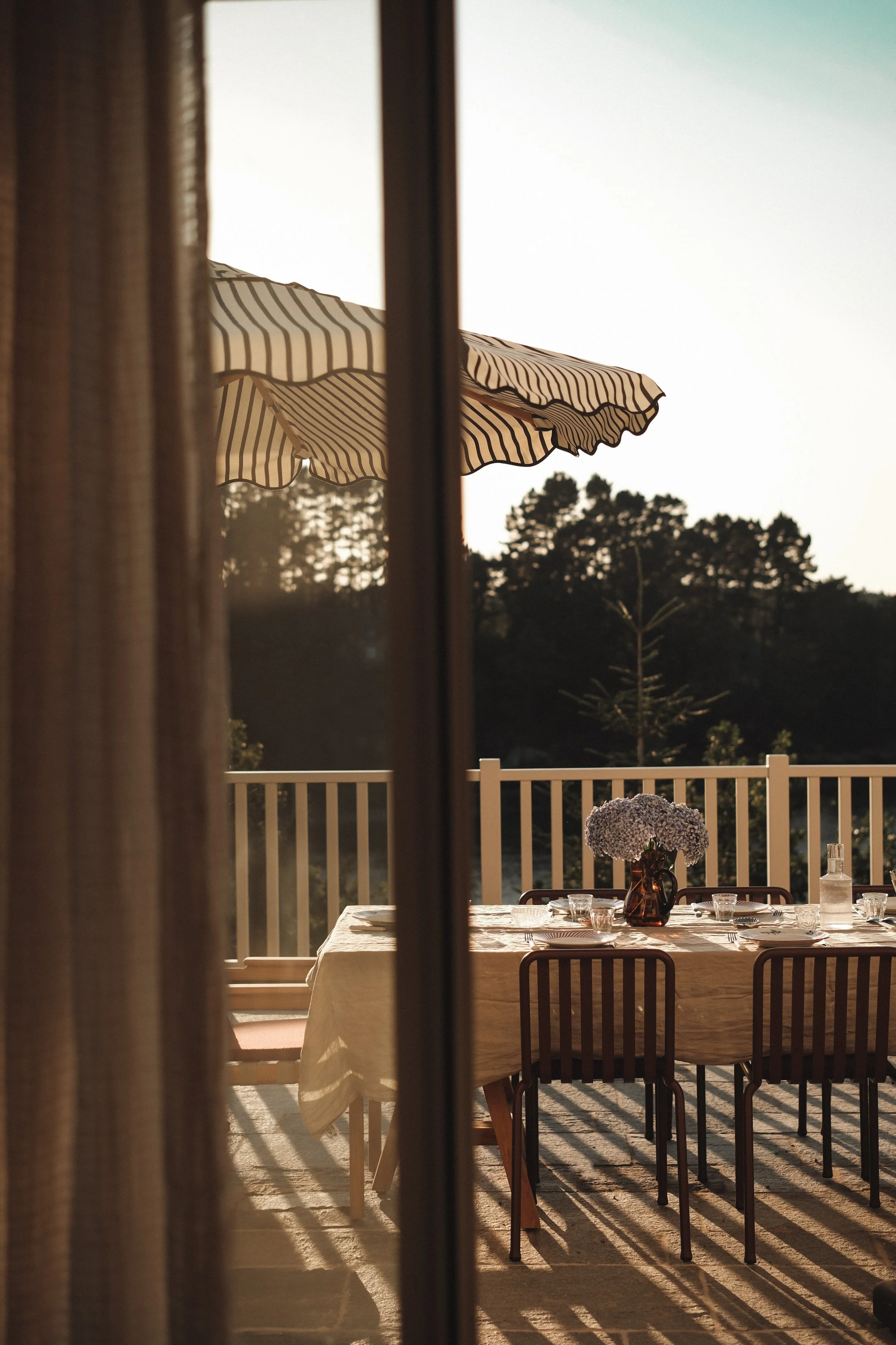 Une table de cuisine extérieure avec un tapis beige, des chaises en bois, des verres, une bouteille d'eau, une carafe de fleurs violettes, une balustrade blanche, un parasol beige à rayures, et des arbres en arrière-plan au coucher du soleil.