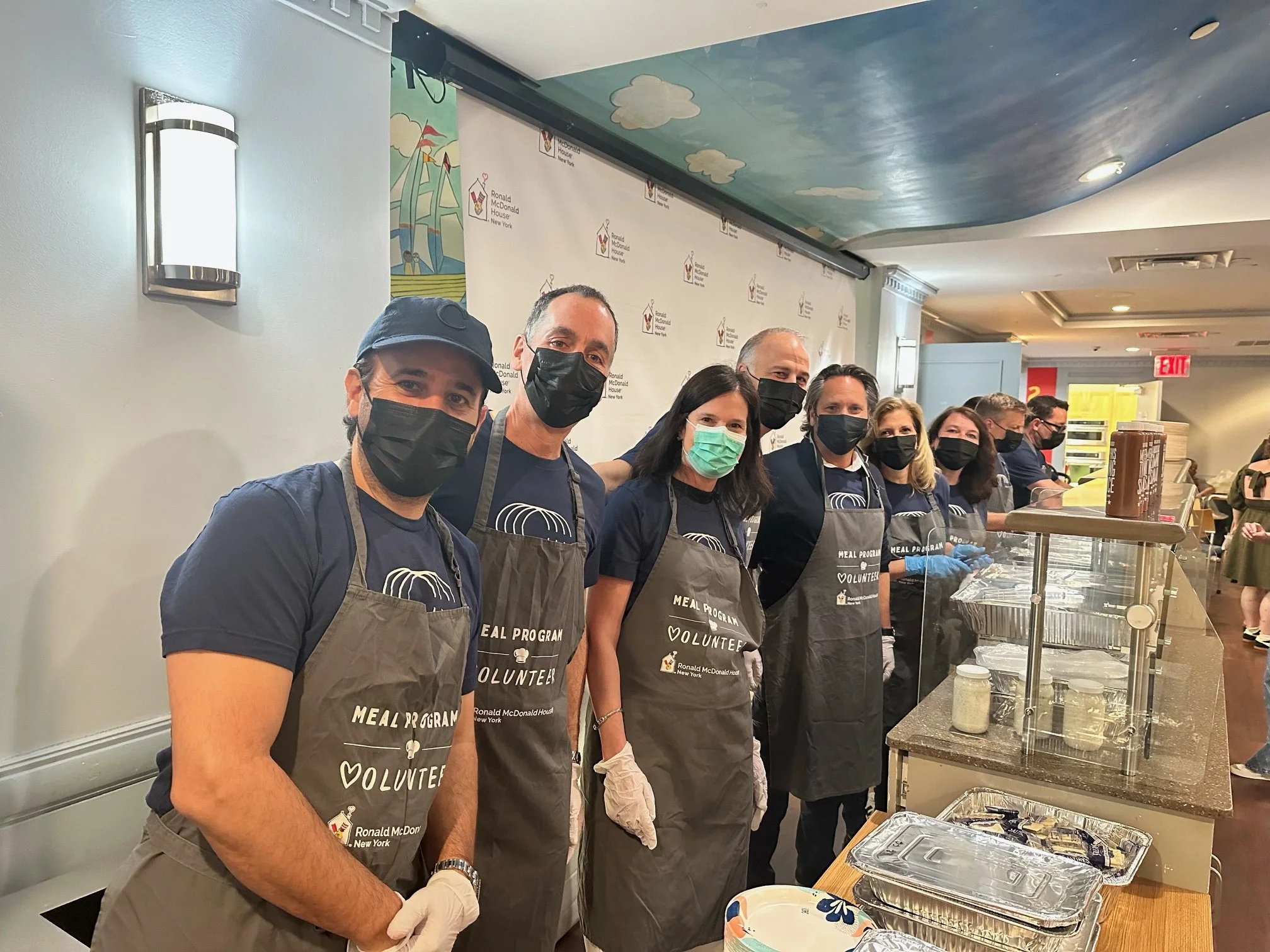 A group of volunteers wearing aprons and masks standing in line at a food service counter at the Ronald McDonald House in New York.