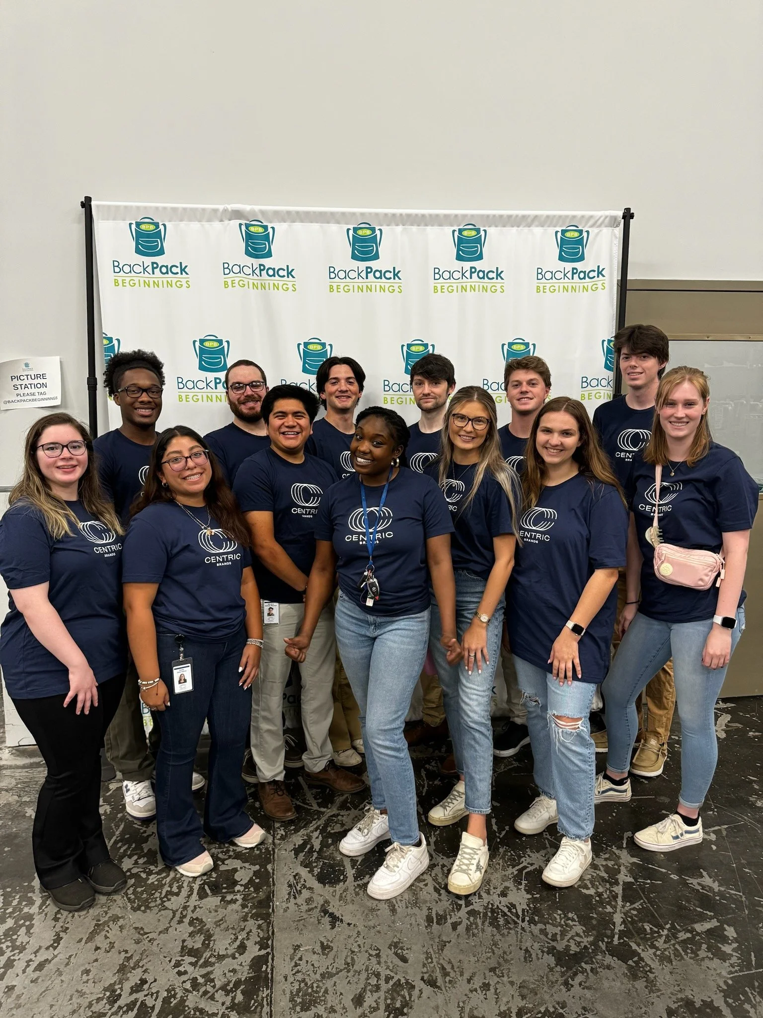 Group of young adults smiling and posing for a photo in front of a BackPack Beginnings banner at an event, all wearing navy blue shirts with the Centric logo.