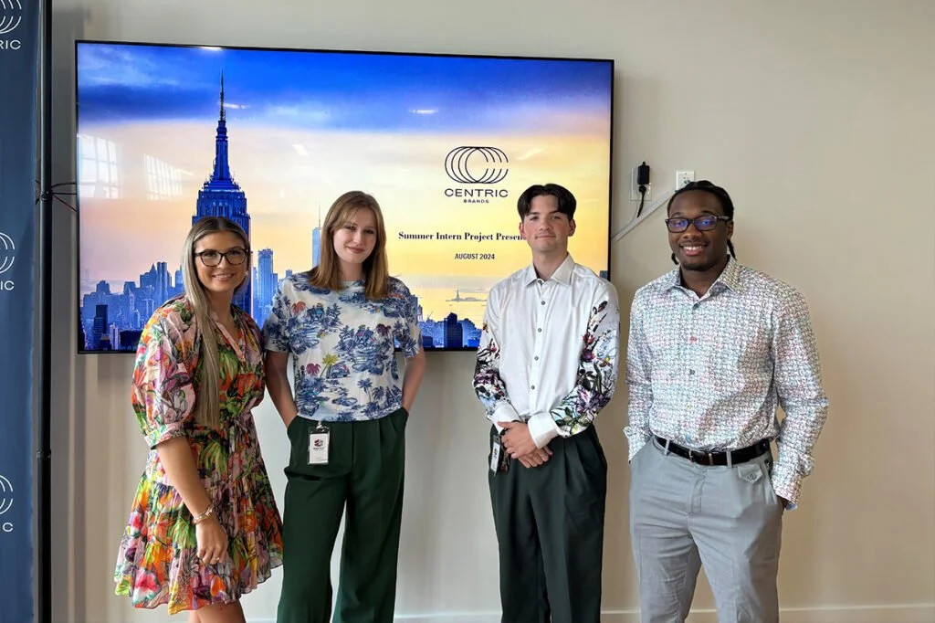 Four young adults standing in front of a large screen displaying the NYC skyline and a logo for Centric Brands, with the text "Summer Intern Project Presentation August 2024".